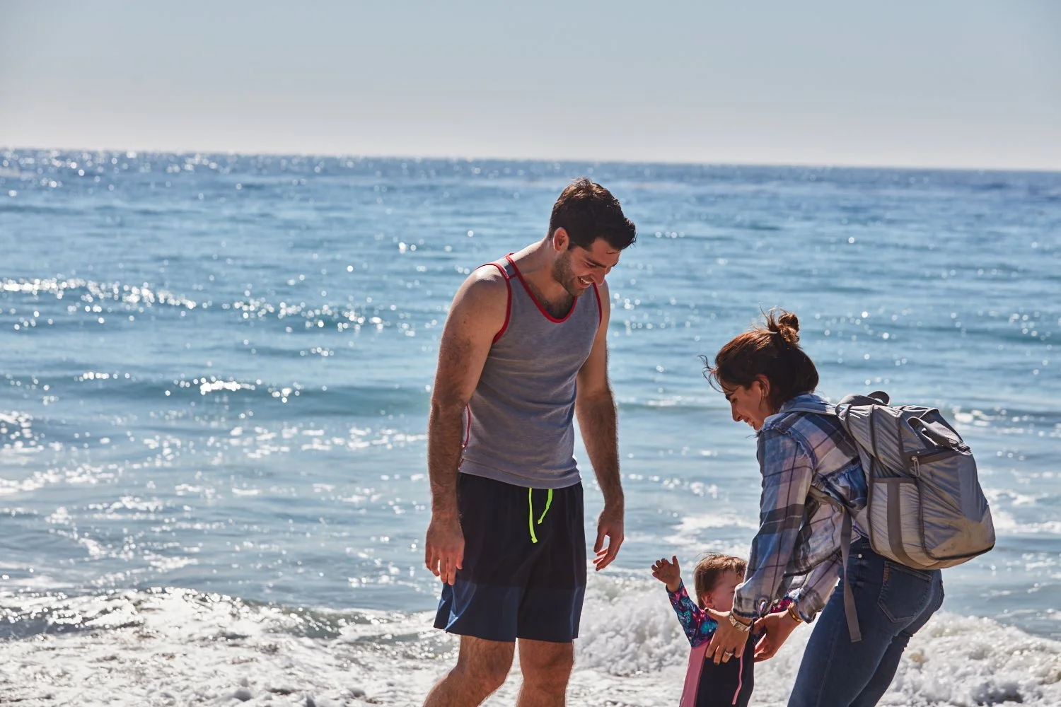 A family at the beach, with a man, woman, and child playing in the surf on a sunny day.