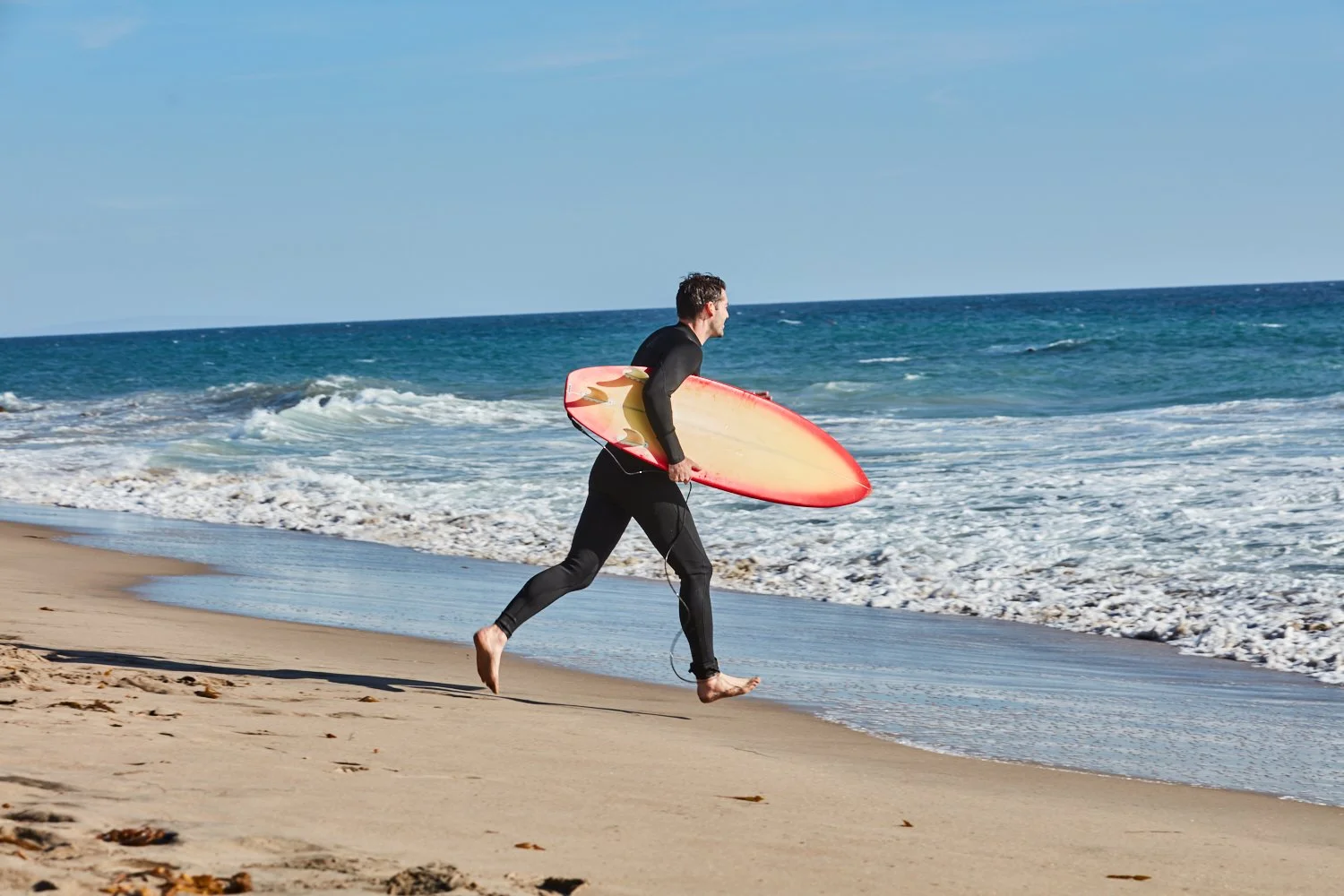 Man in black wetsuit carrying a surfboard on a sandy beach with ocean waves in the background.