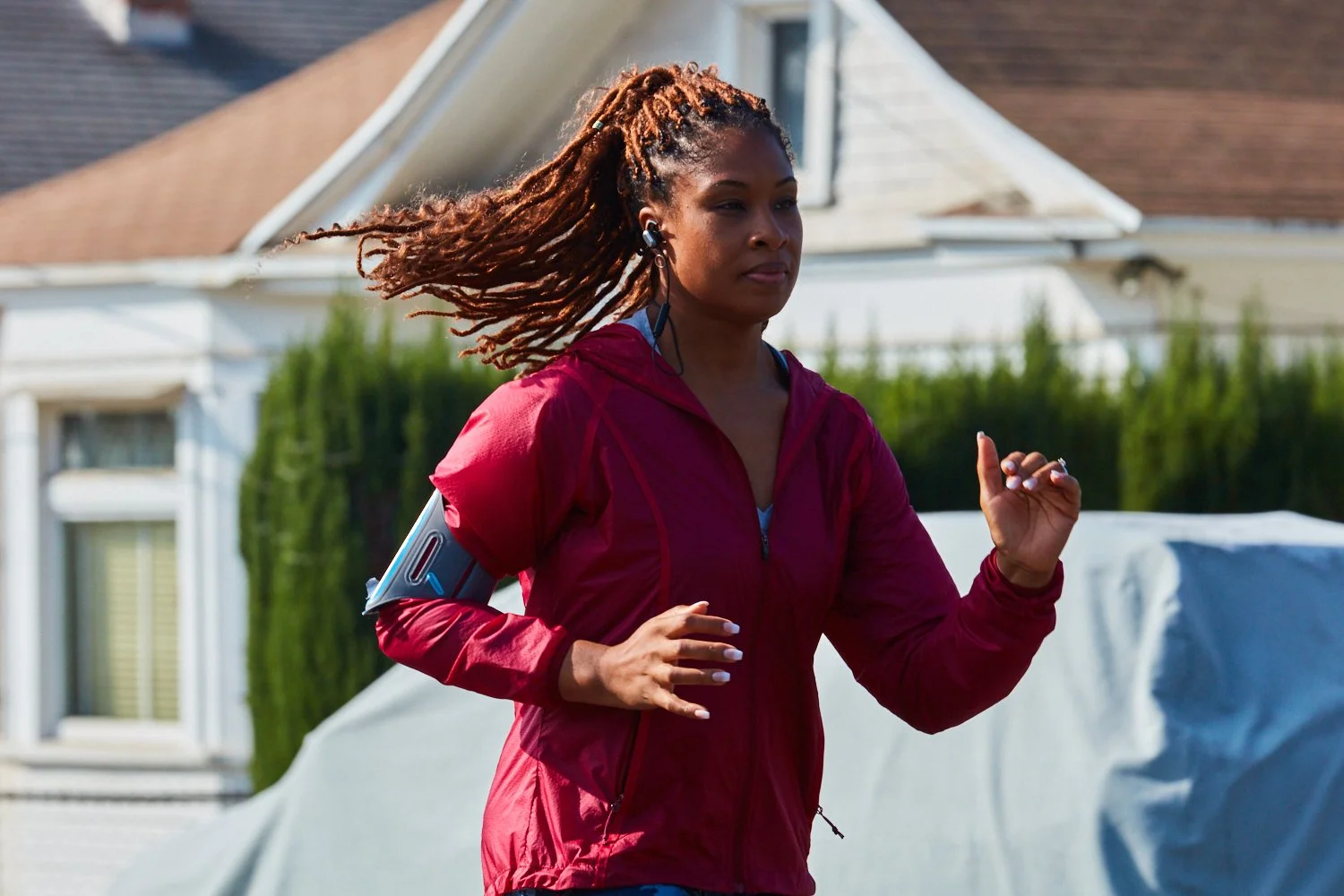 Woman with long dreadlocks running outdoors in a residential area, wearing a red jacket and earphones.