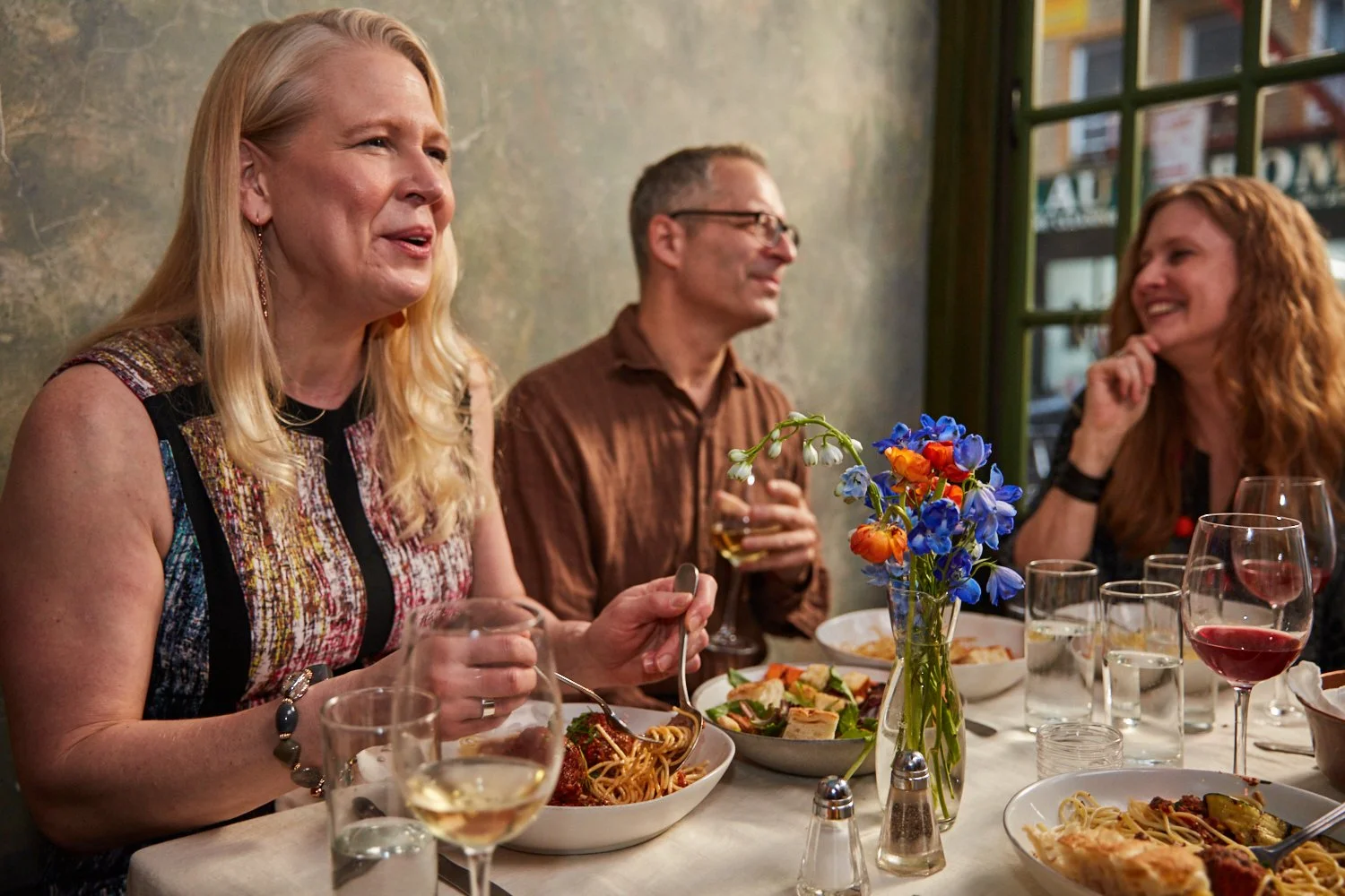 Three people sitting at a restaurant table enjoying a meal and drinks, with a vase of colorful flowers in the center.