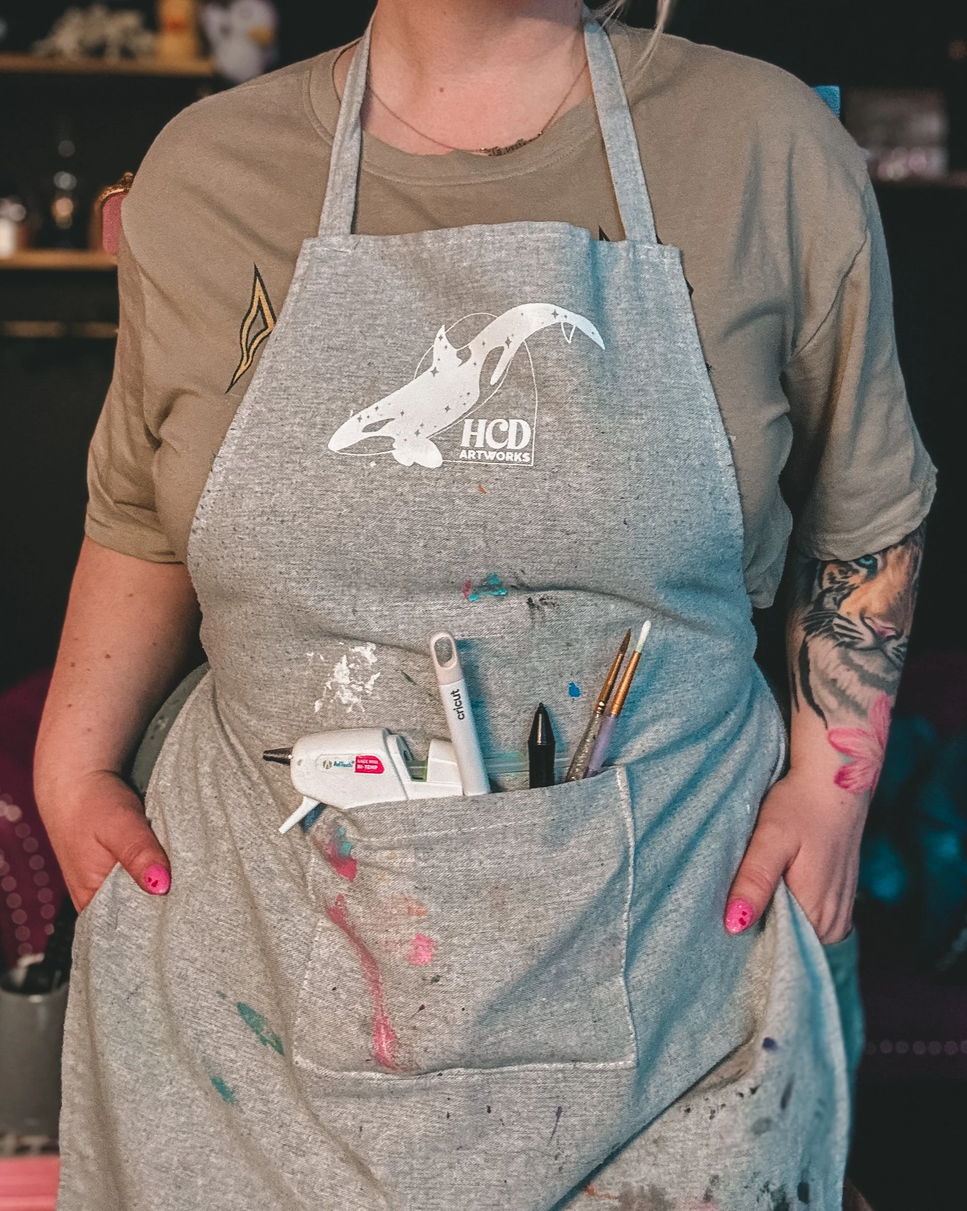 Heather "HCD" wearing a grey artist apron with a white silhouette HCD Artworks logo in the upper centre. A glue gun, Cricut weeder, digital pen, and paintbrushes sit in the front square pocket.