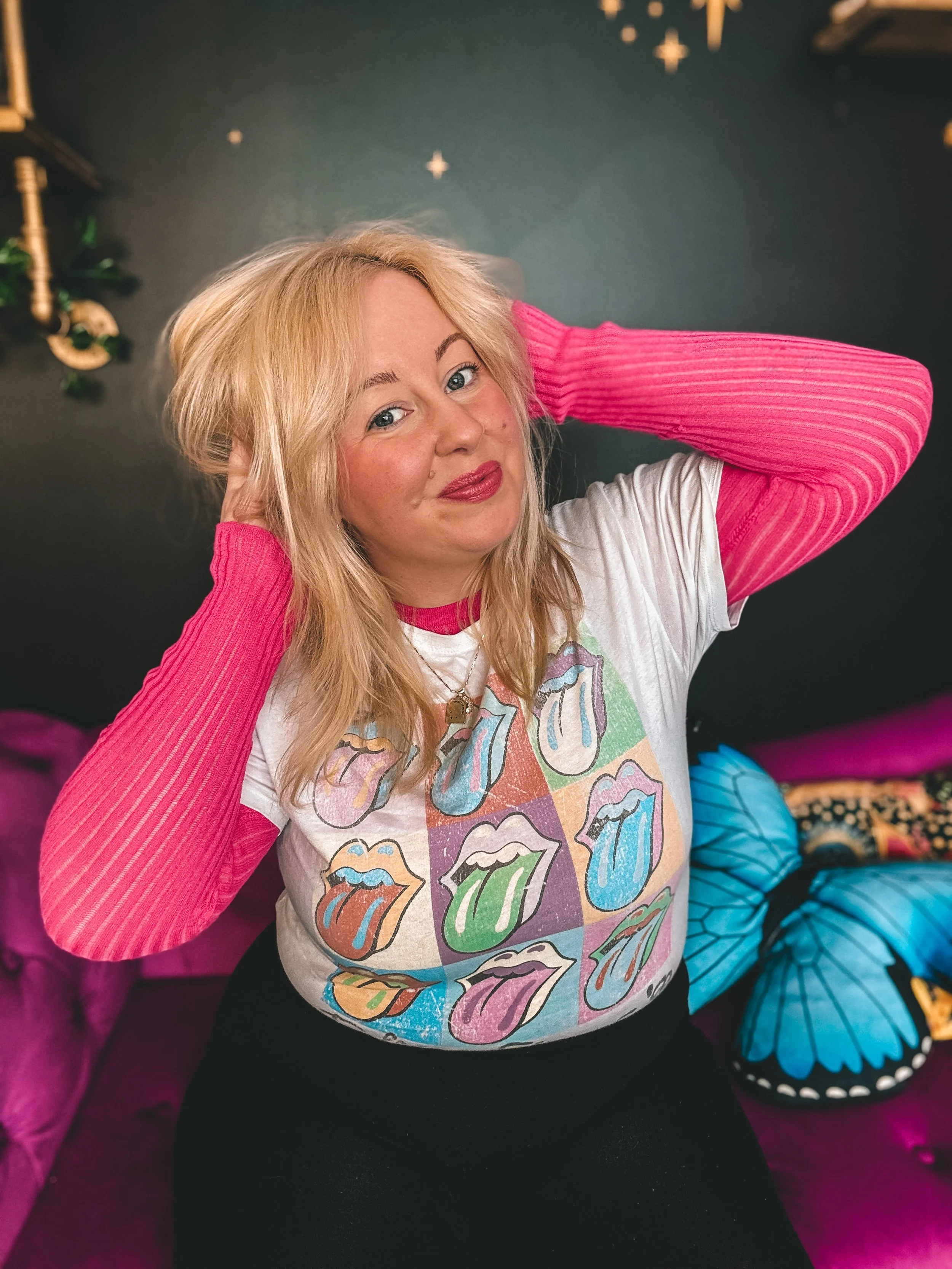 A photo of Heather. She has blonde hair, wearing a white t-shirt with colourful Rolling Stones logo graphics, over a pink long-sleeve mesh shirt, & black pants. She's sitting on a pink/purple sofa, with her hands tousling her hair, smiling.