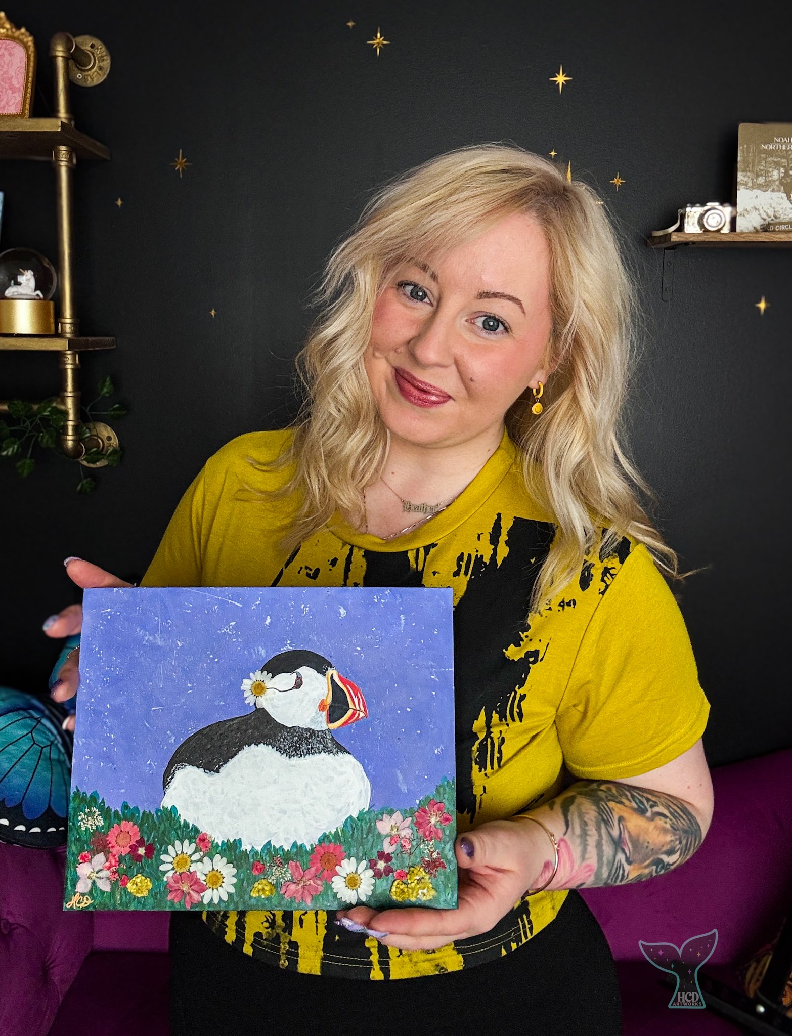 Woman with blonde hair, tattoos on her arm, wearing a yellow T-shirt, holding a painting of a puffin bird surrounded by colorful flowers, standing in front of a black wall with gold star decals.
