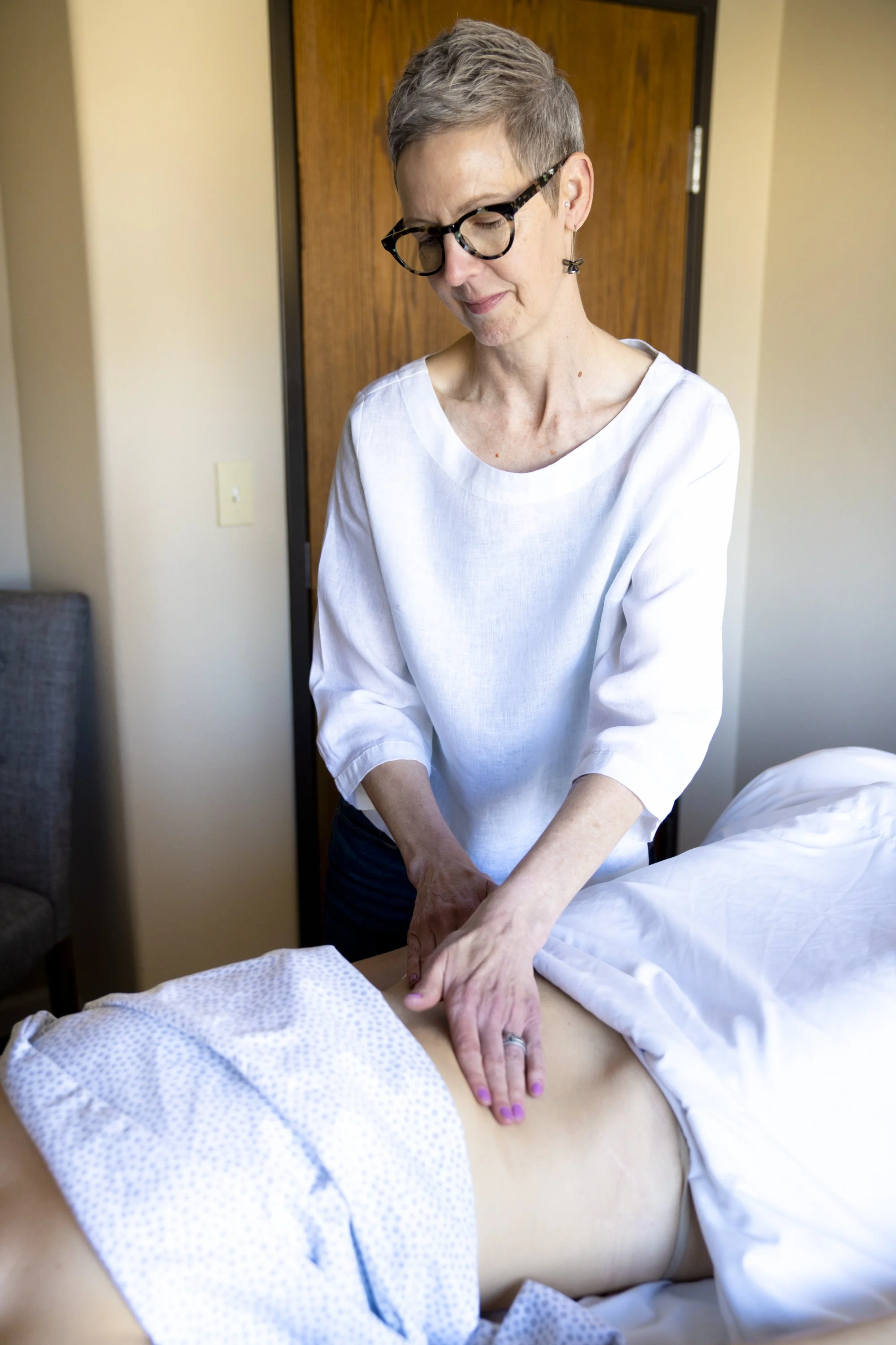 A woman with short gray hair and glasses performing a massage on a patient's back in a clinical setting.