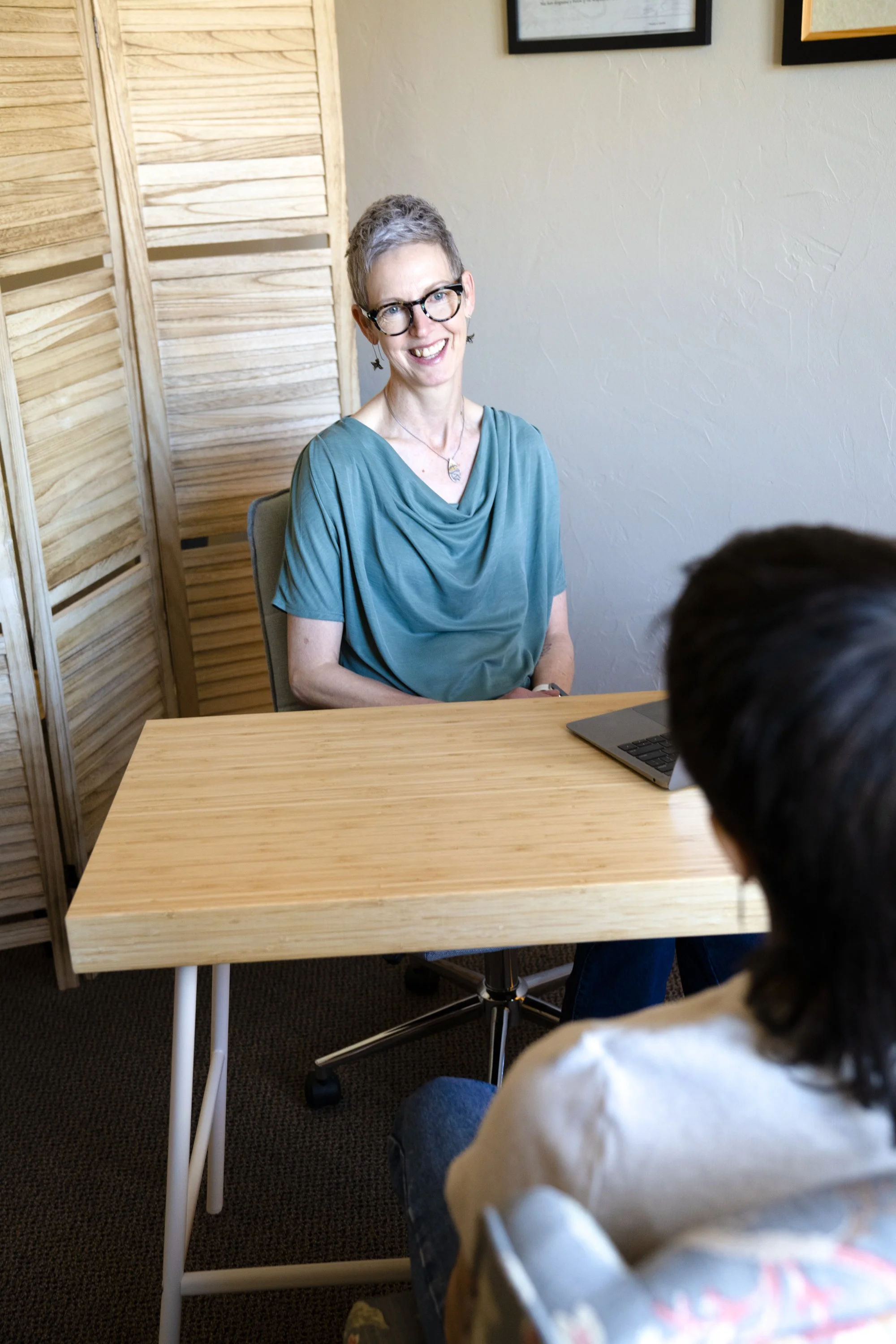 A woman with short gray hair, glasses, and earrings sitting at a wooden table, smiling, with a person in the foreground.