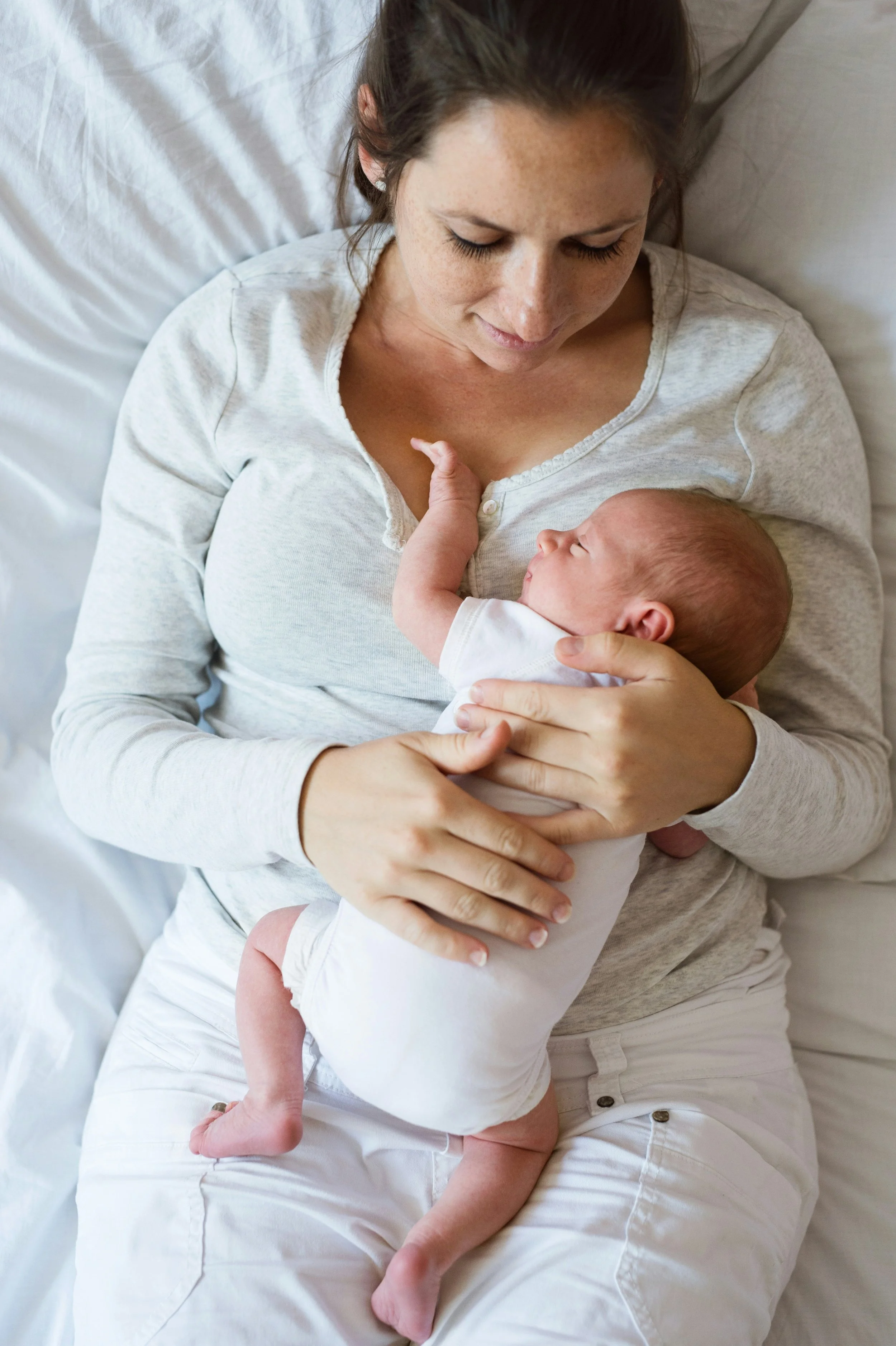 A woman with dark hair and freckles, dressed in a light-colored shirt and pants, lying on a bed and breastfeeding a baby, who is dressed in a white onesie.