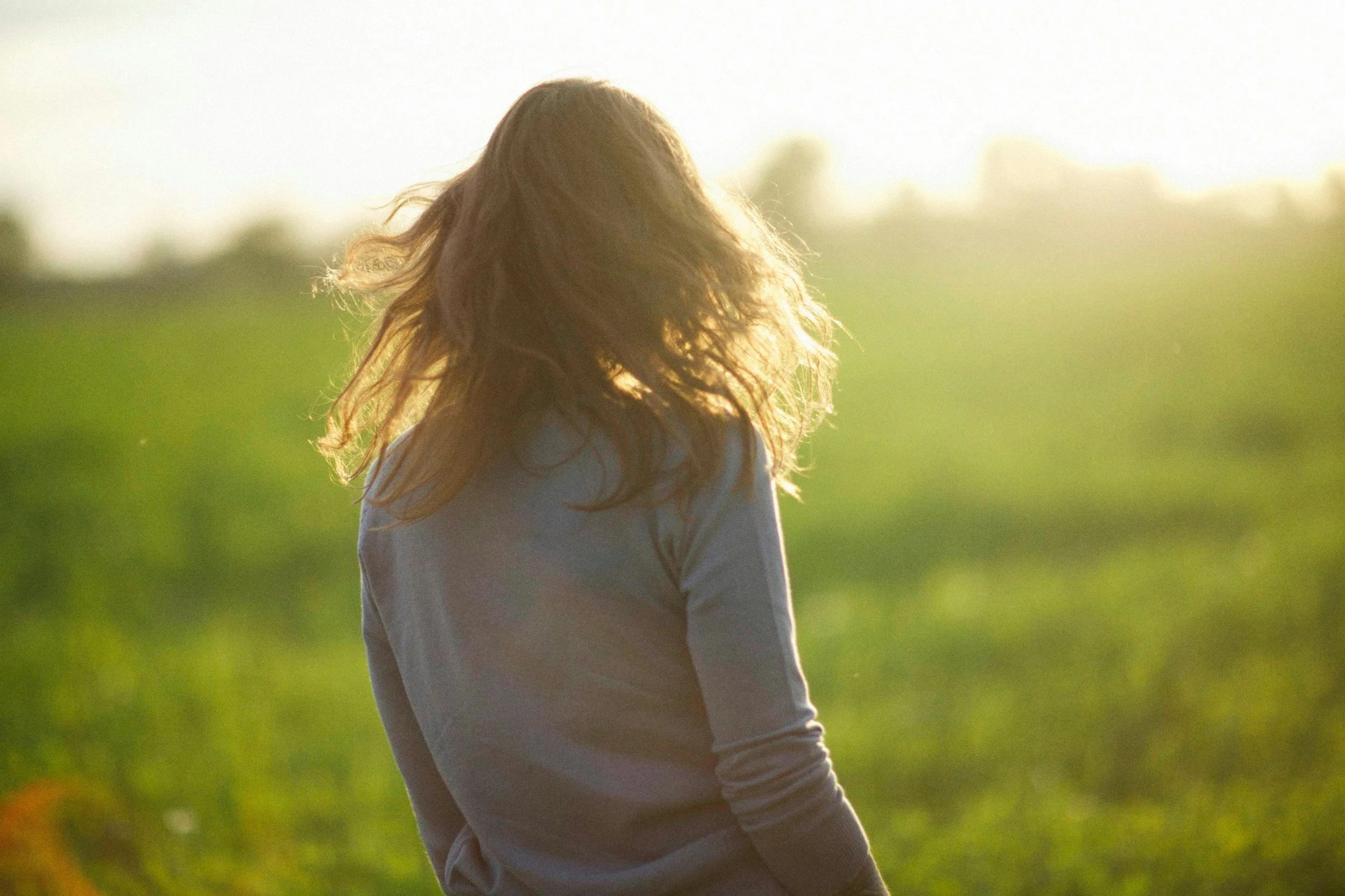A woman with long, wavy hair stands in a field during sunset, facing away from the camera.