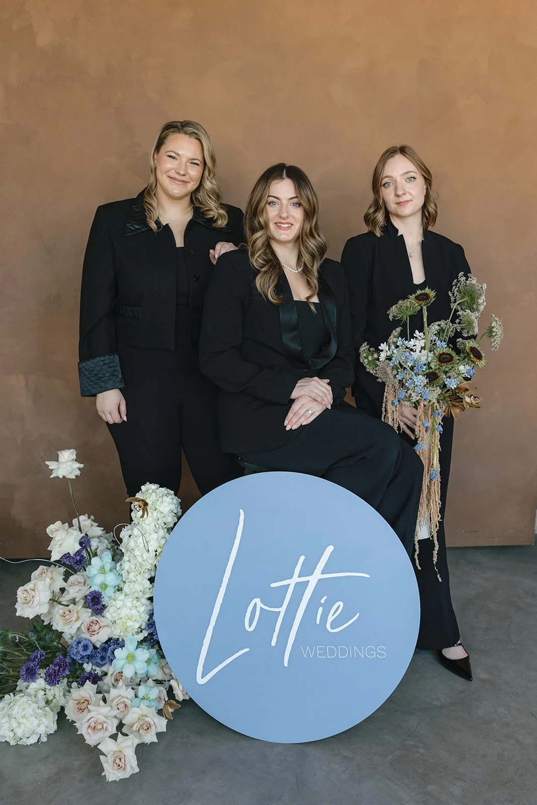 Three women in black formal attire posing with floral arrangements and a blue sign that says "Lottie Weddings" in cursive text, standing against a brown backdrop.