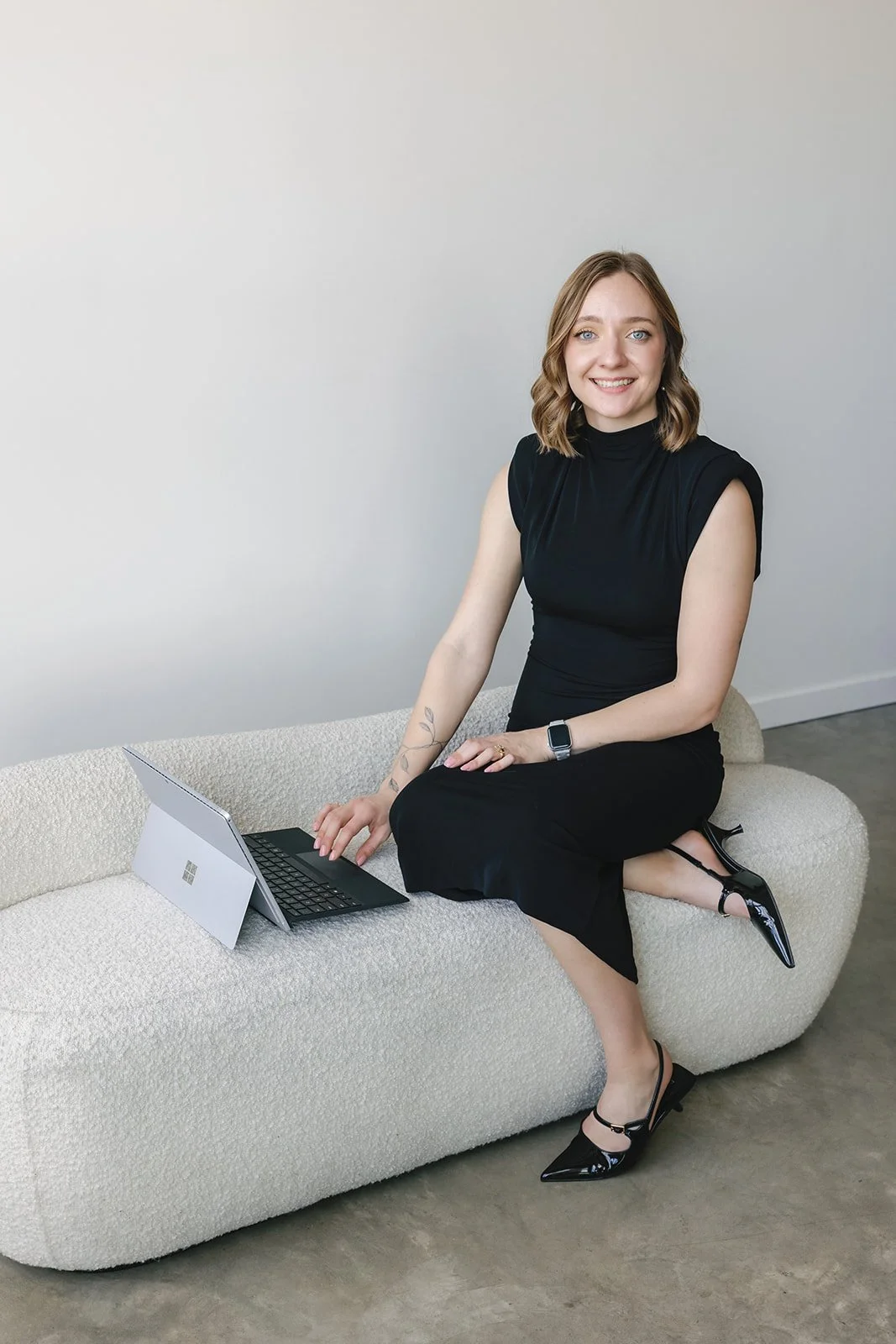 A young woman with wavy blonde hair sitting on a light-colored textured sofa in a black sleeveless top and black skirt, smiling at the camera with a Microsoft Surface tablet and keyboard in front of her.