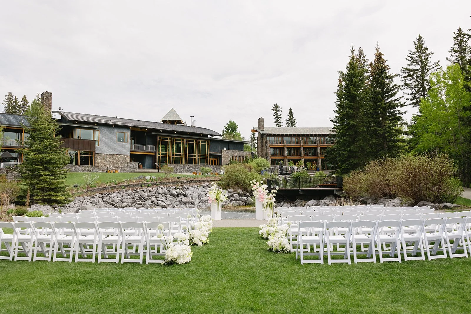 Outdoor wedding ceremony at Azuridge Estate Hotel in Priddis, Alberta, featuring white chairs, lush white and pink floral arrangements, and a scenic woodland backdrop.