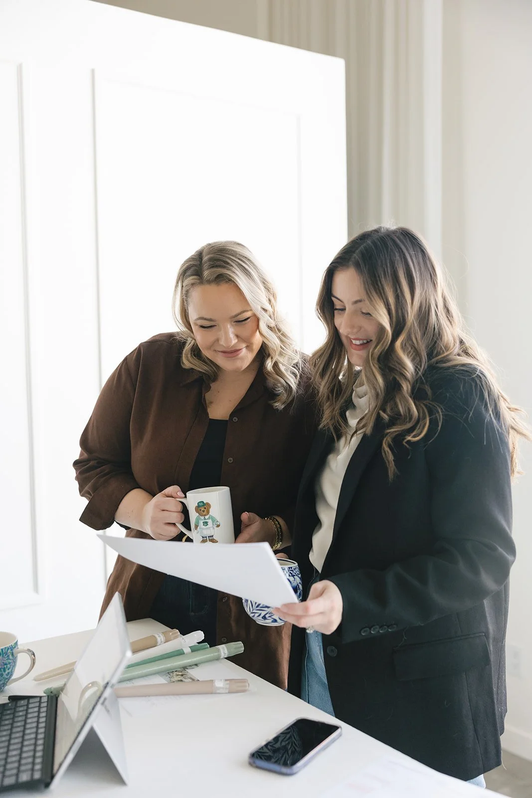 Two women looking at papers together in an office, one holds a mug with a bear illustration, the other holds a blue and white mug, on a desk with a laptop, phone, and rolled-up papers.
