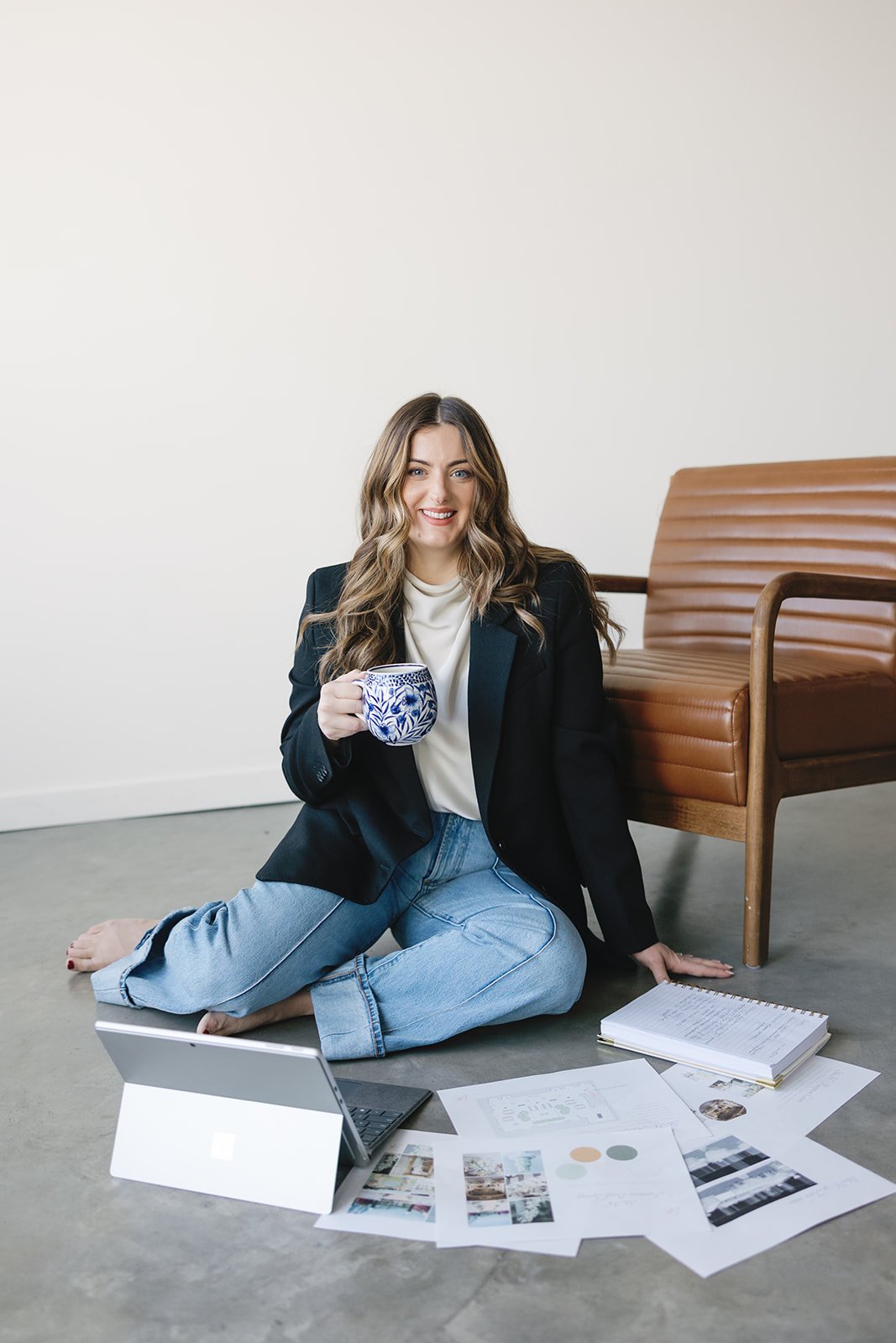 A woman sitting on the floor with a laptop, holding a mug, surrounded by papers, notebooks, and printed images, with a wooden chair beside her.