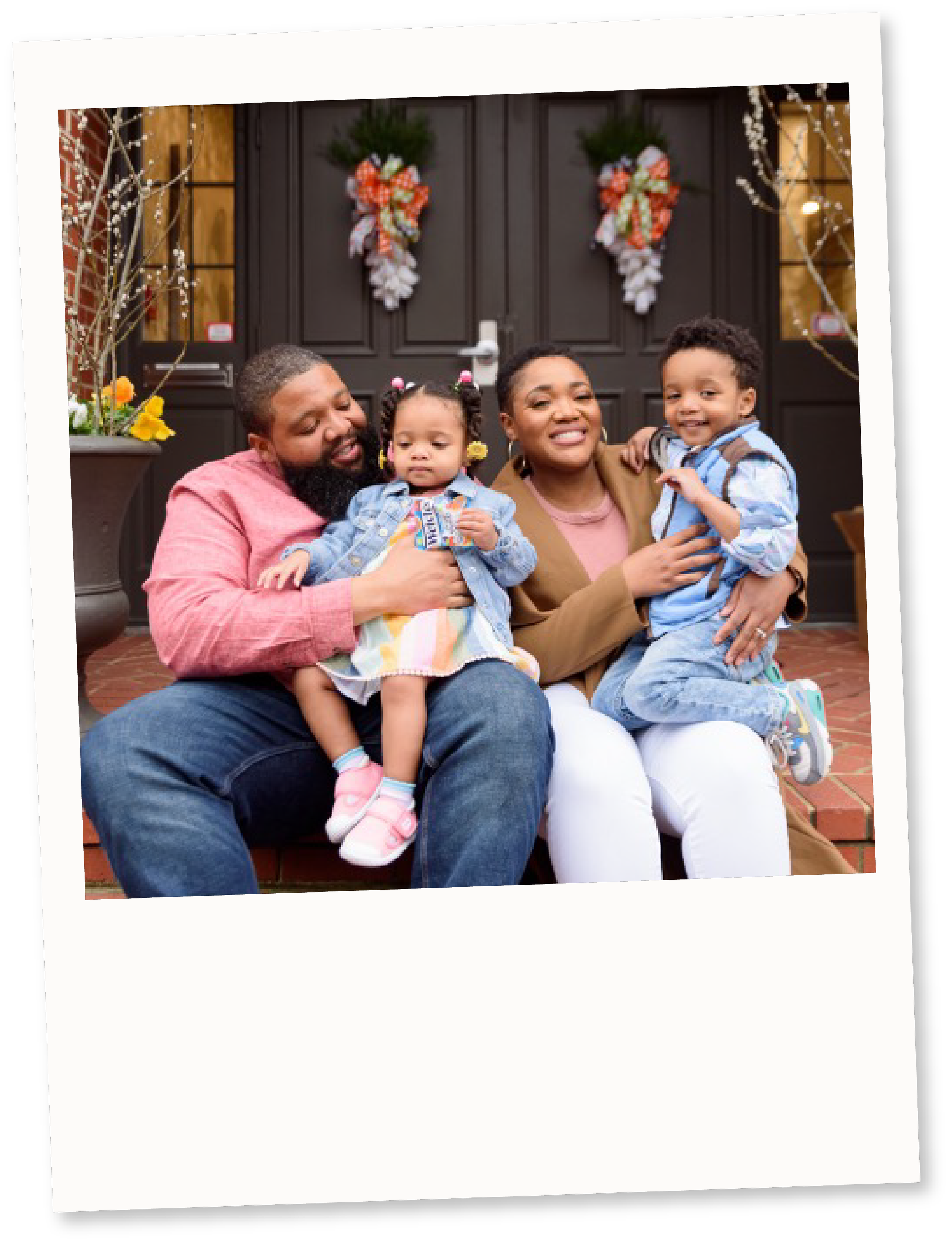 Family of four sitting on steps in front of a decorated door, smiling and holding children, with Christmas wreaths and decorations.