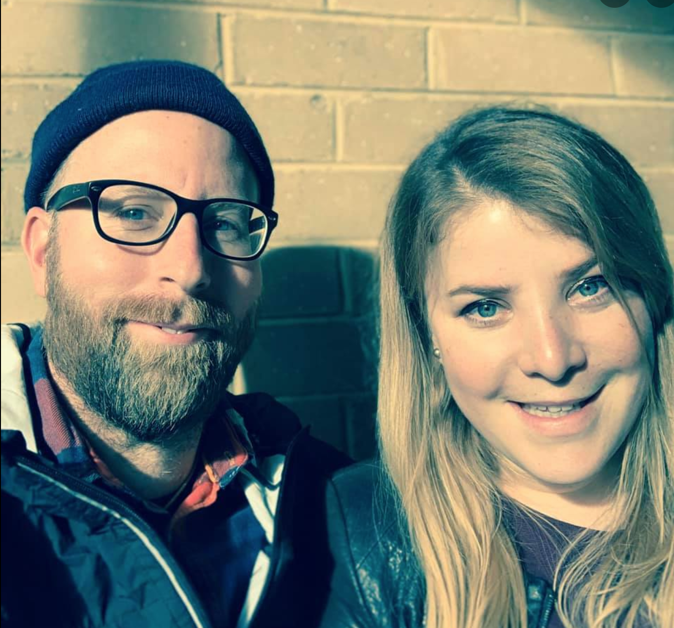 A smiling man with glasses, a beanie, and a beard taking a selfie with a woman with long blond hair.