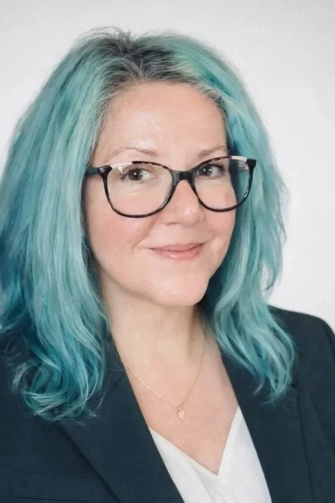 A woman with shoulder-length blue hair, wearing glasses, a black blazer, and a gold necklace, smiling at the camera against a plain background.