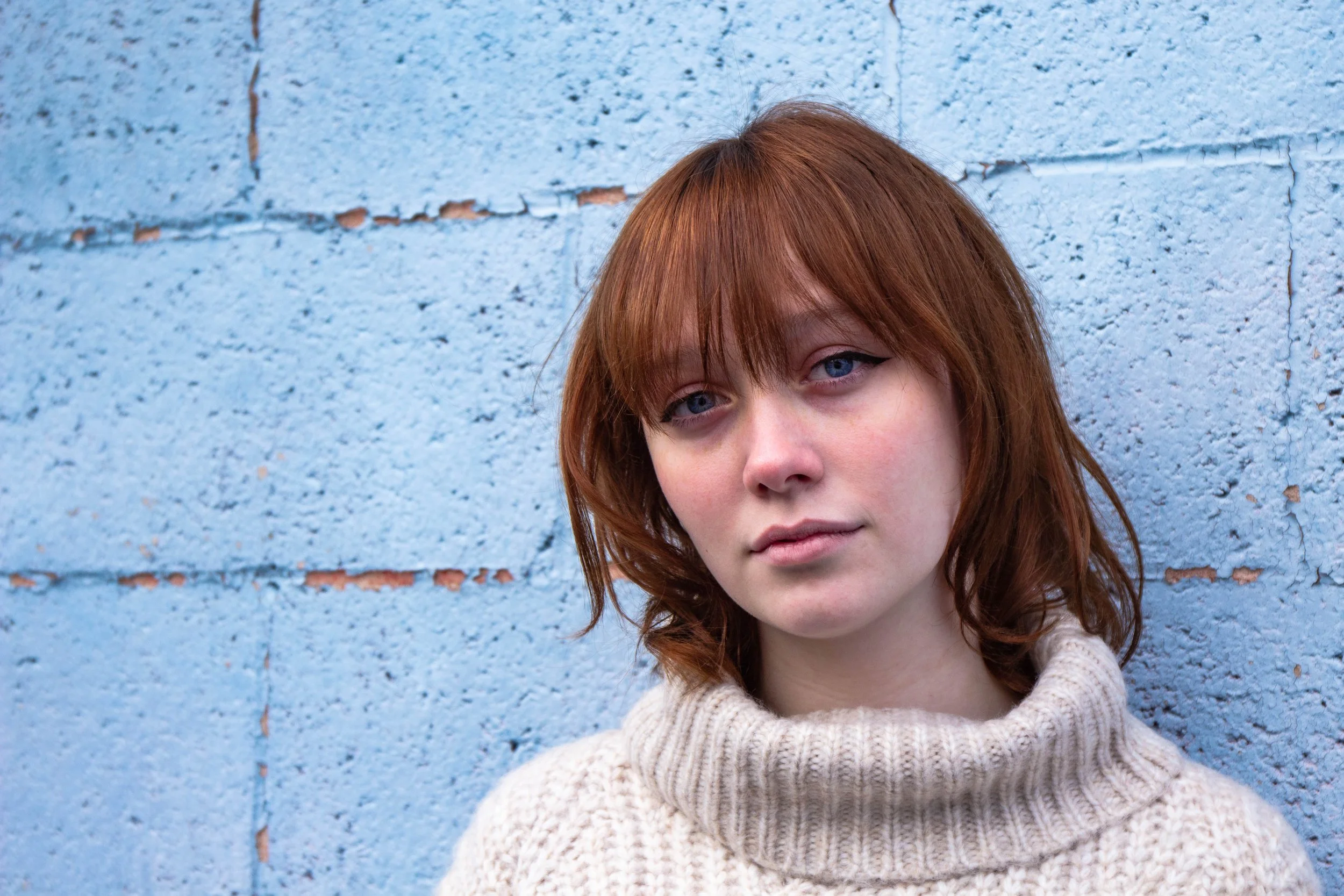 Portrait photography by Gayle Bower: woman with red hair and blue eyes standing against a textured blue brick wall, editorial-style natural light photo
