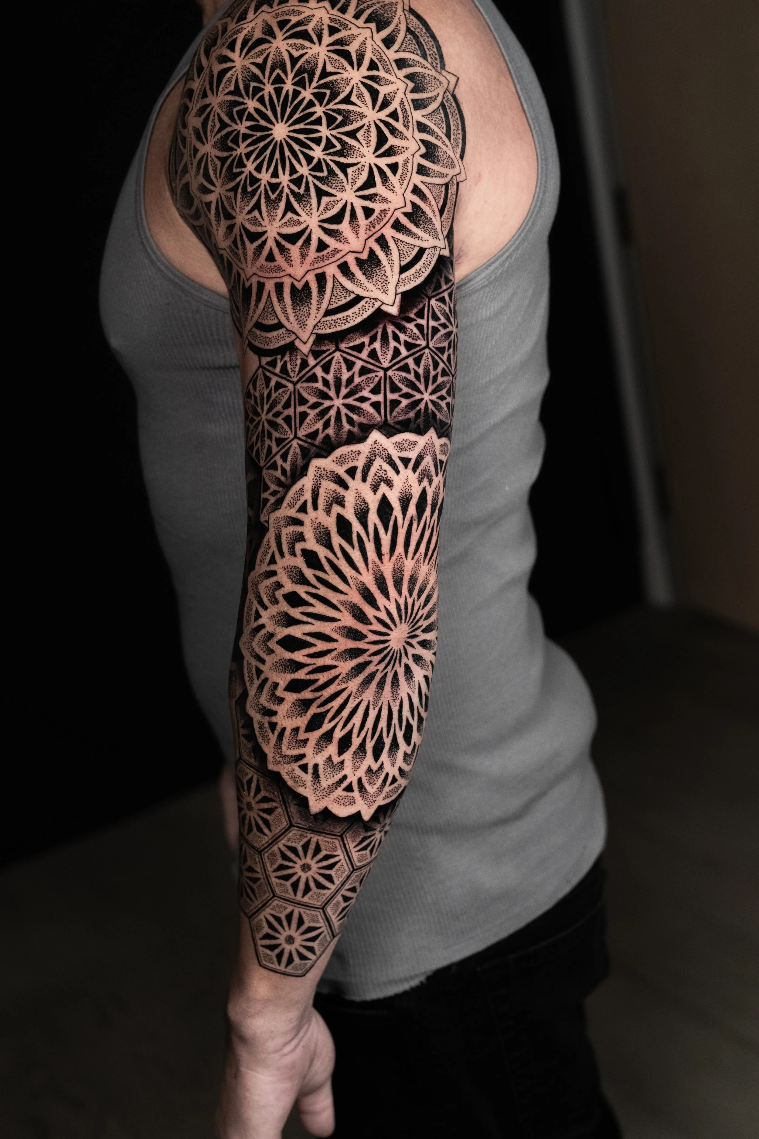 A person's arm featuring a large, intricate black and natural skin tone geometric tattoo in mandala and hexagon patterns.