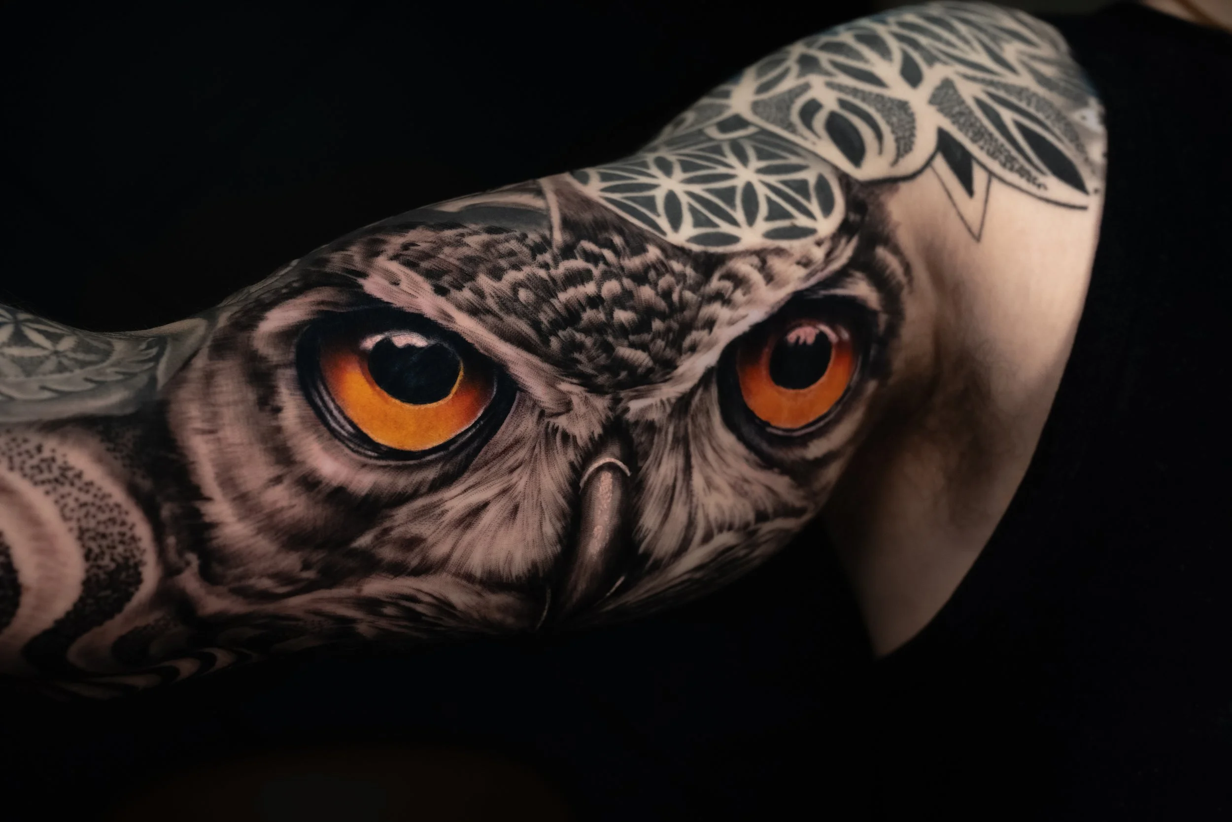 Close-up of a tattooed arm with an owl's face and orange eyes, against a black background.