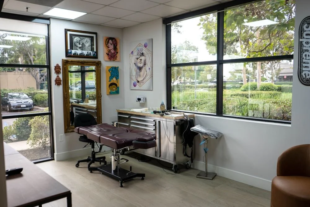 Tattoo studio examination room with tattoo chair, large window, artwork on walls, and medical supplies.