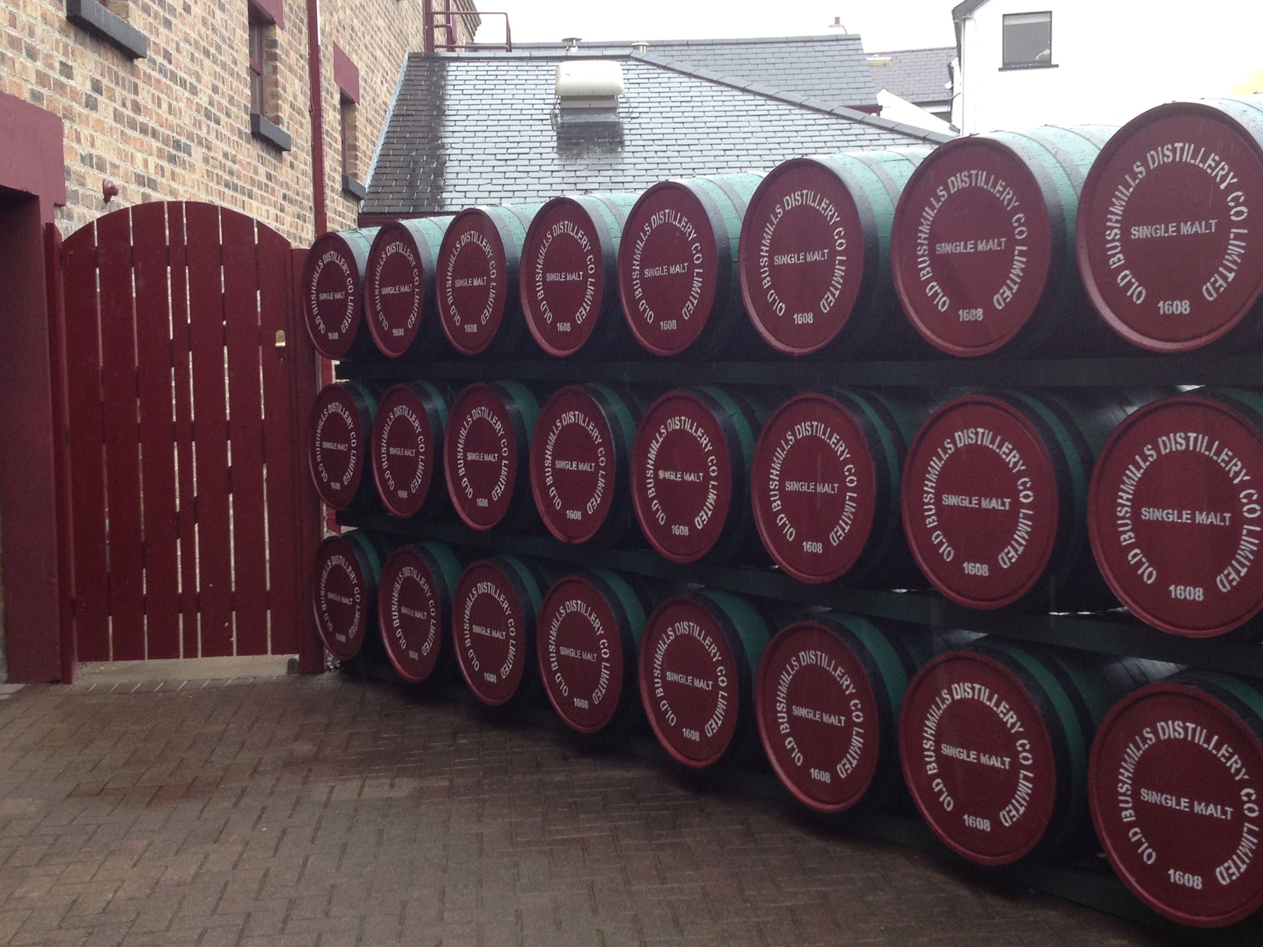 Stacked barrels labeled 'Oild Bushmills Distillery Co Limited 1608 Single Malt' next to a red wooden gate in an alley with brick and brick-paved ground.
