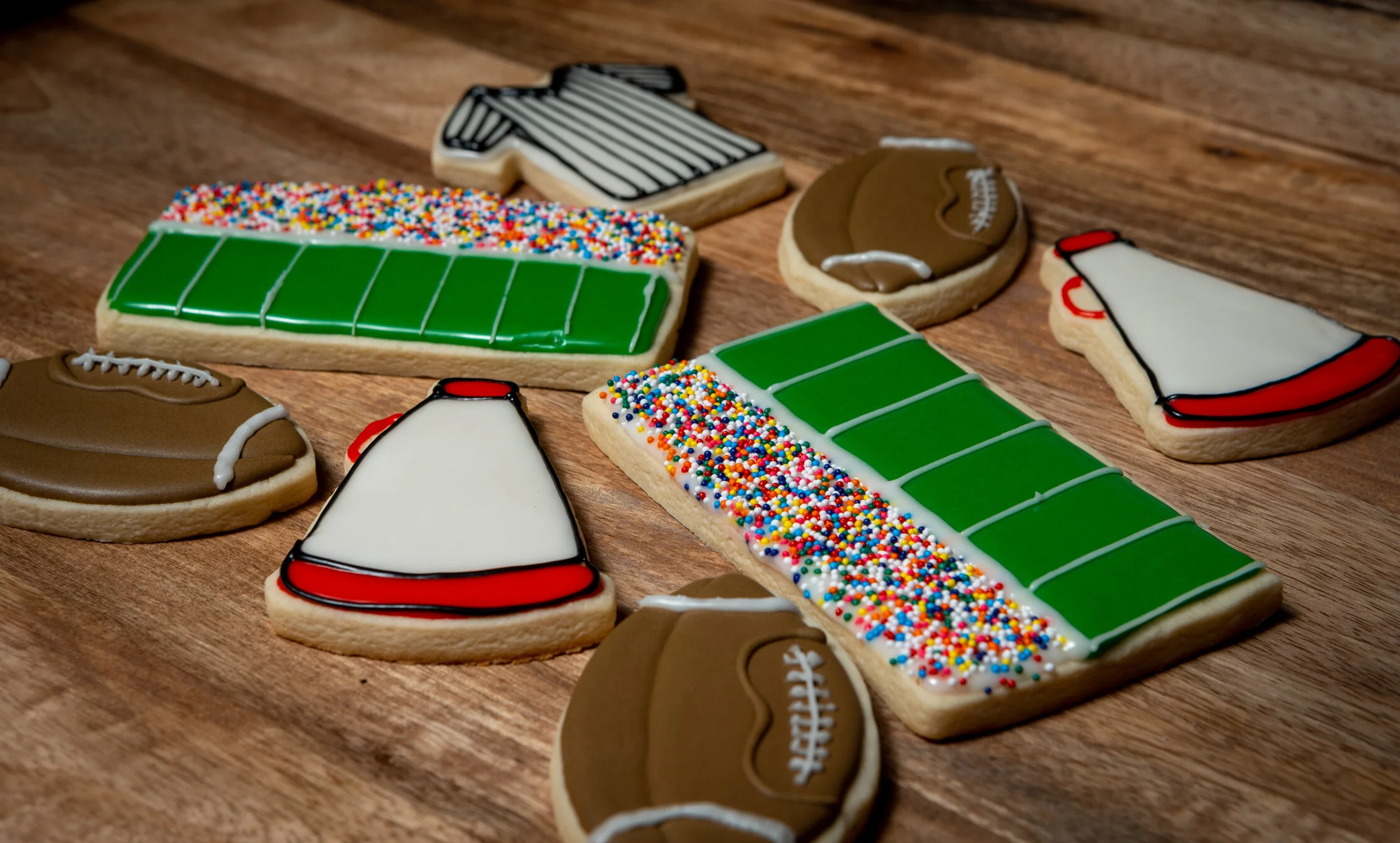 Football-themed decorated sugar cookies on a wooden surface, shaped and decorated like footballs and football fields with colorful icing and sprinkles.