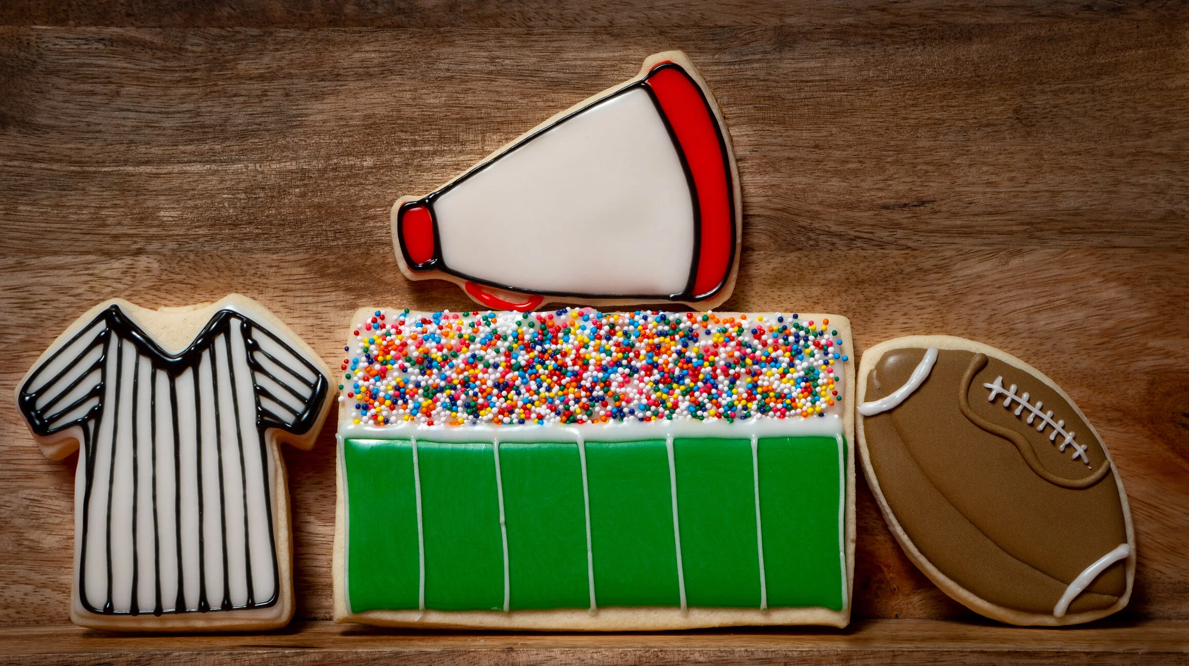 Sports-themed decorated cookies including a football jersey, a megaphone, a football, and a section of a green football field with rainbow sprinkles, all on a wooden surface.