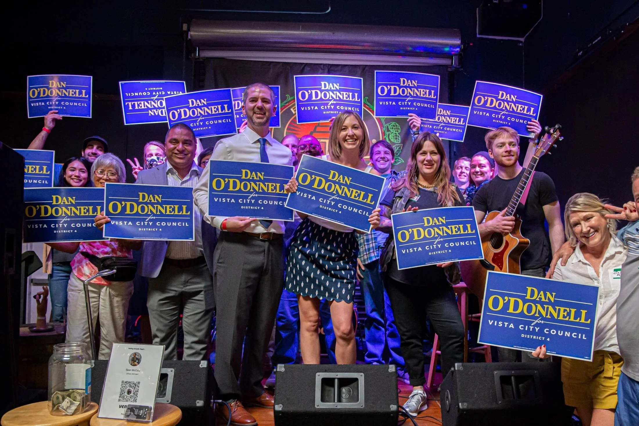 Group of people on stage holding campaign signs for Dan O'Donnell for Vista City Council District 4, smiling and celebrating, with some making peace signs, and one person playing a guitar.