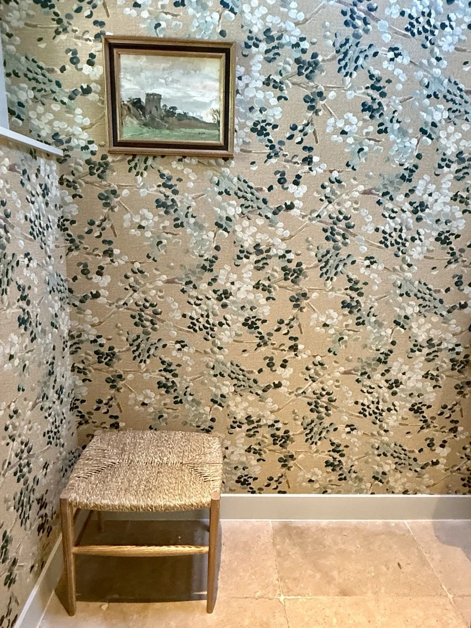Een kleine houten stoel met een gevlochten zitting, geplaatst in een hoek van een kamer met bloempatriottapete. Boven de stoel hangt een kader met een schilderij van een oud kasteel in een landschap.