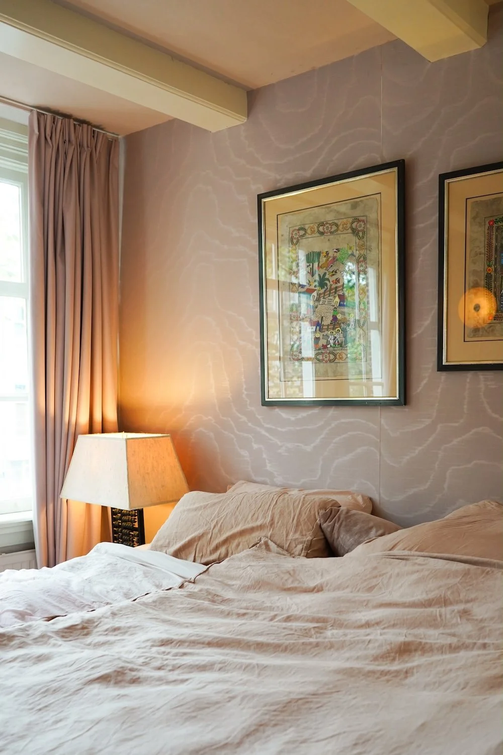 Een slaapkamer met een bed, beige lakens en kussens, een tafelkleed en schilderijen aan de muur, en een raam met beige gordijnen dat natuurlijk licht binnenlaat.