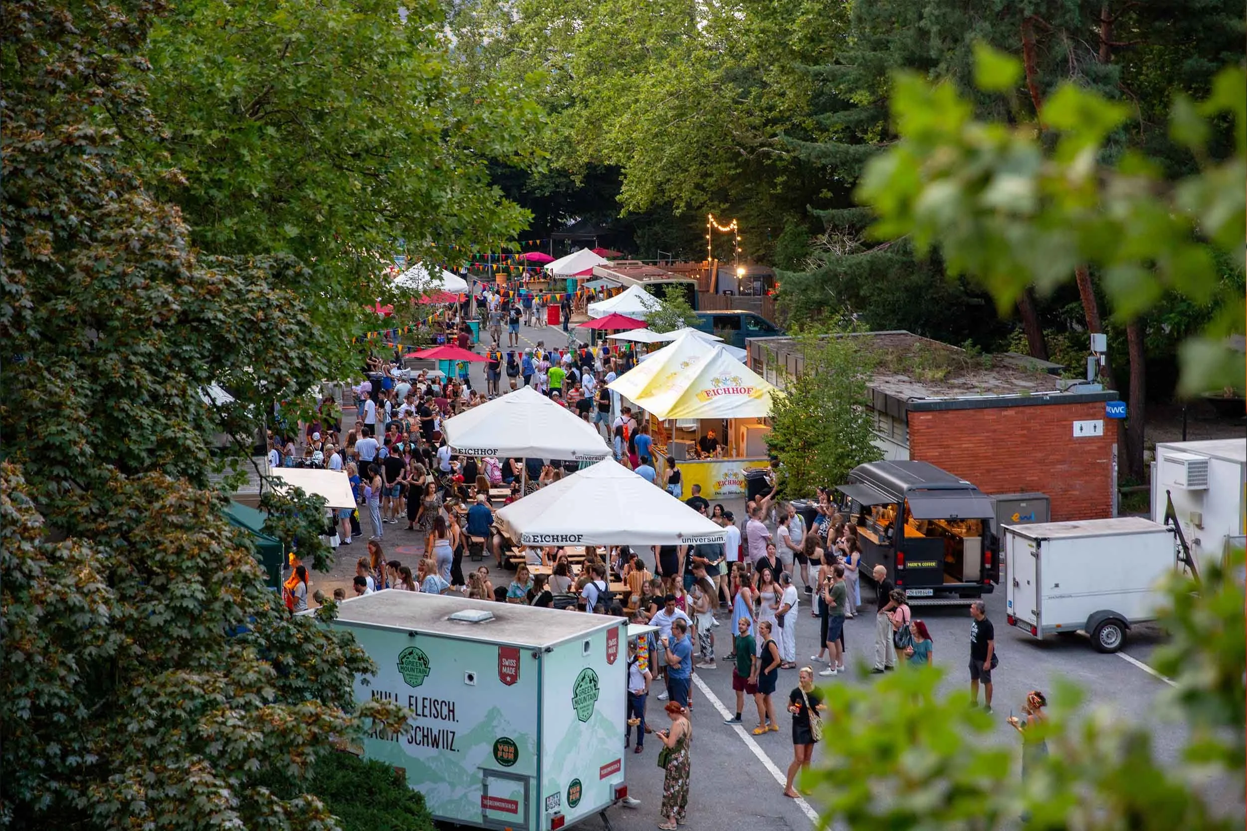 Blick über das Festivalgelände mit Foodtrucks, Sonnenschirmen und Publikum am Inseliquai