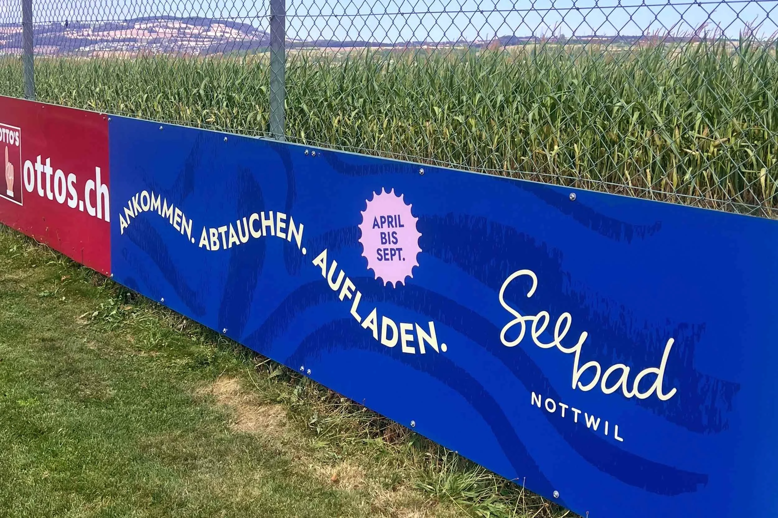 Blaue Bandenwerbung des Seebad Nottwil mit Logo und Slogan «Ankommen, abtauchen, aufladen» am Zaun des lokalen Fussballfelds