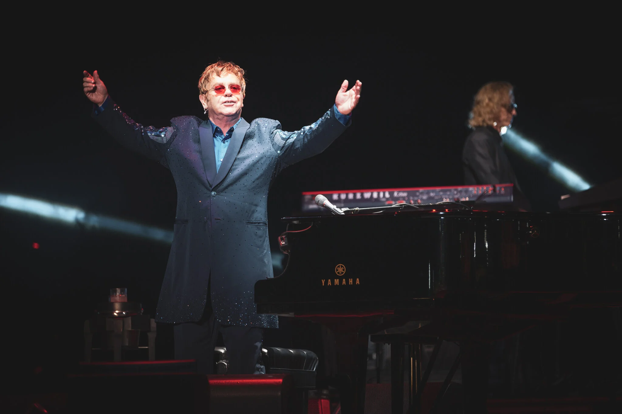 Elton John steht auf einer Bühne neben einem schwarzen Yamaha Klavier, mit ausgestreckten Armen. Elton John trägt einen dunklen, mit Glitzersteinchen besetzten Mantel und rote Sonnenbrille.