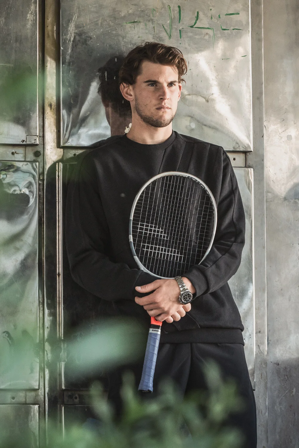 Dominic Thiem steht an einer metallischen Wand, hält einen Tennisracket, trägt schwarze Kleidung und eine Armbanduhr, schaut ernst.