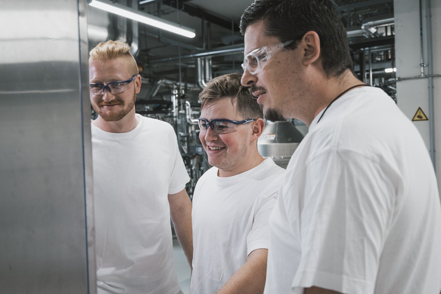 Drei Männer in weißen Laborkitteln und Schutzbrillen, die in einem technischen Kontrollraum arbeiten.