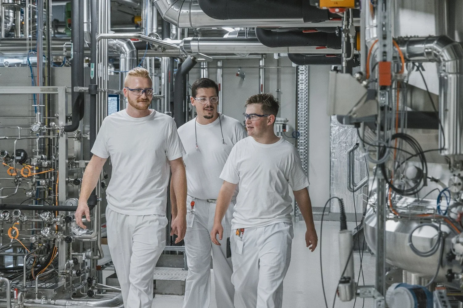 Drei Männer in weißen Arbeitskleidung und Schutzbrillen gehen durch eine technische Anlagenhalle mit vielen Rohren und Maschinen.