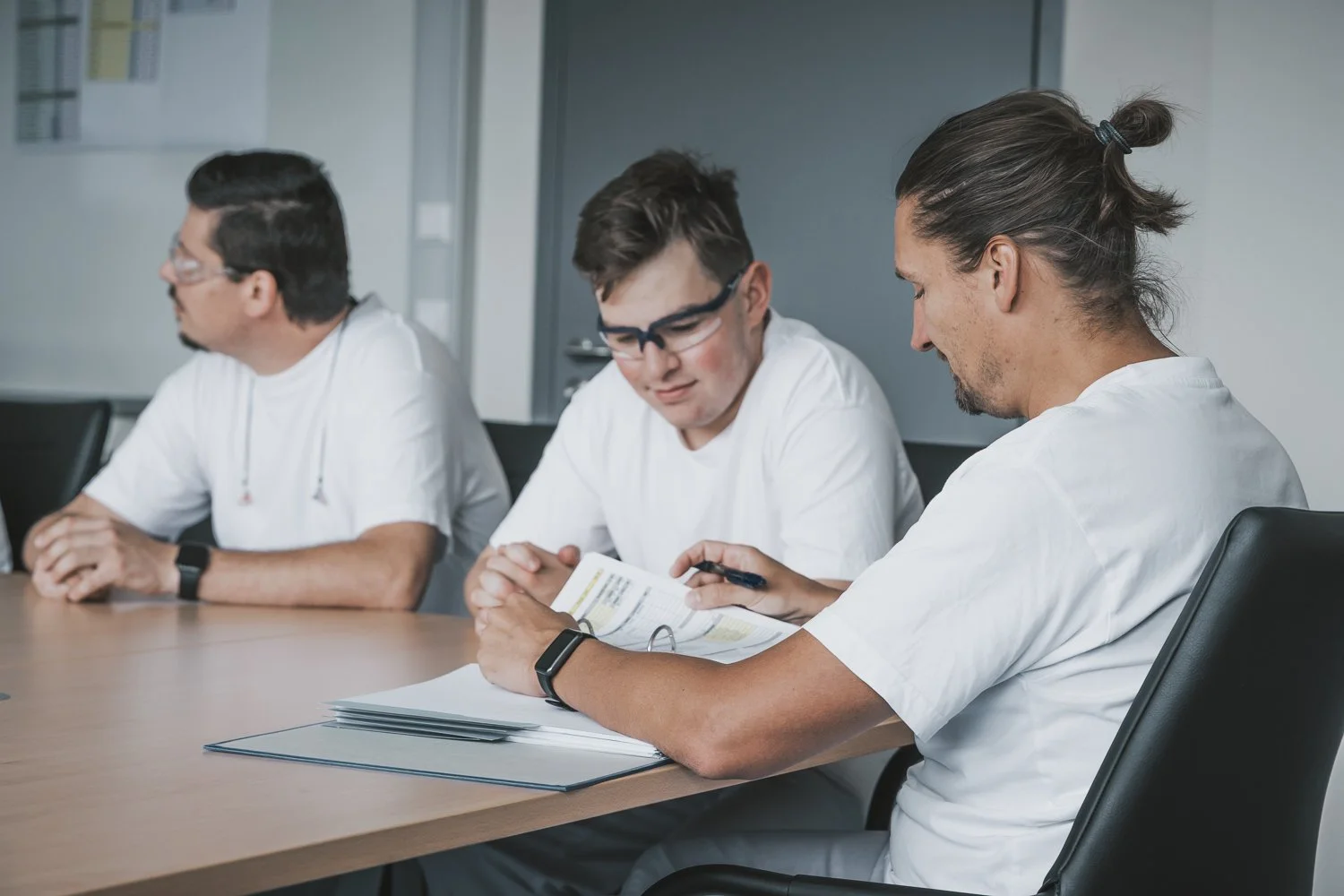 Drei Männer in weißen medizinischen Kitteln sitzen an einem Tisch in einem Besprechungsraum. Der Mann im Vordergrund liest in einem Dokument, der Mann in der Mitte schaut zu, und der Mann im Hintergrund sieht nach links. Es sind Unterlagen und ein St