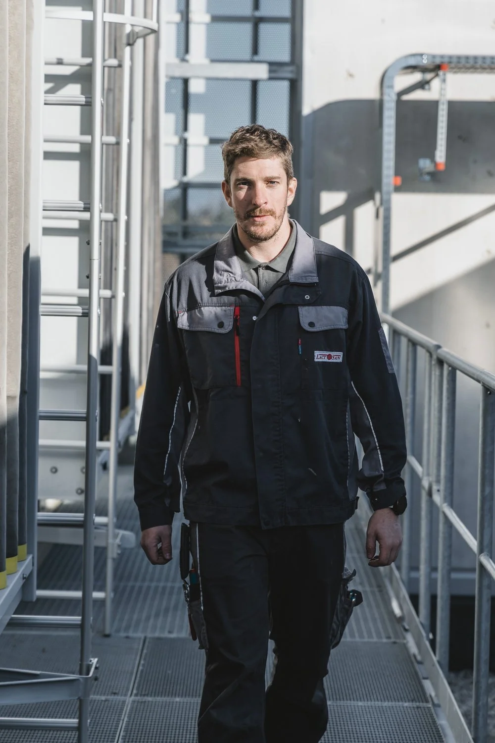 Mann in dunkler Arbeitshandschuhen und schwarzer Jacke läuft auf einer Metallplattform in einem Industriegebiet.