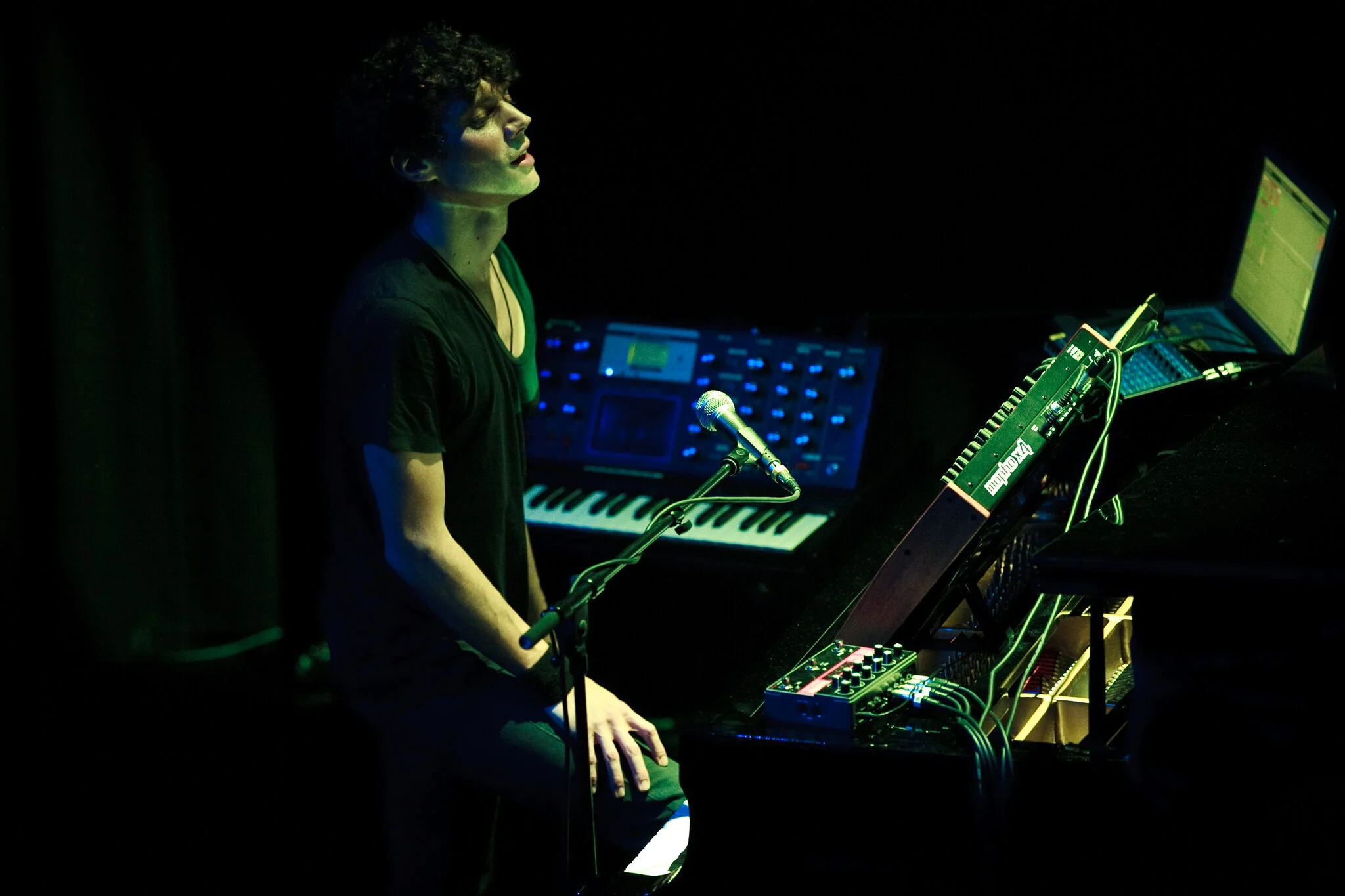 Ein junger Mann sitzt an einem Mischpult auf einer dunklen Bühne, umgeben von elektronischen Musikinstrumenten und einem Laptop, während er singt oder spricht.