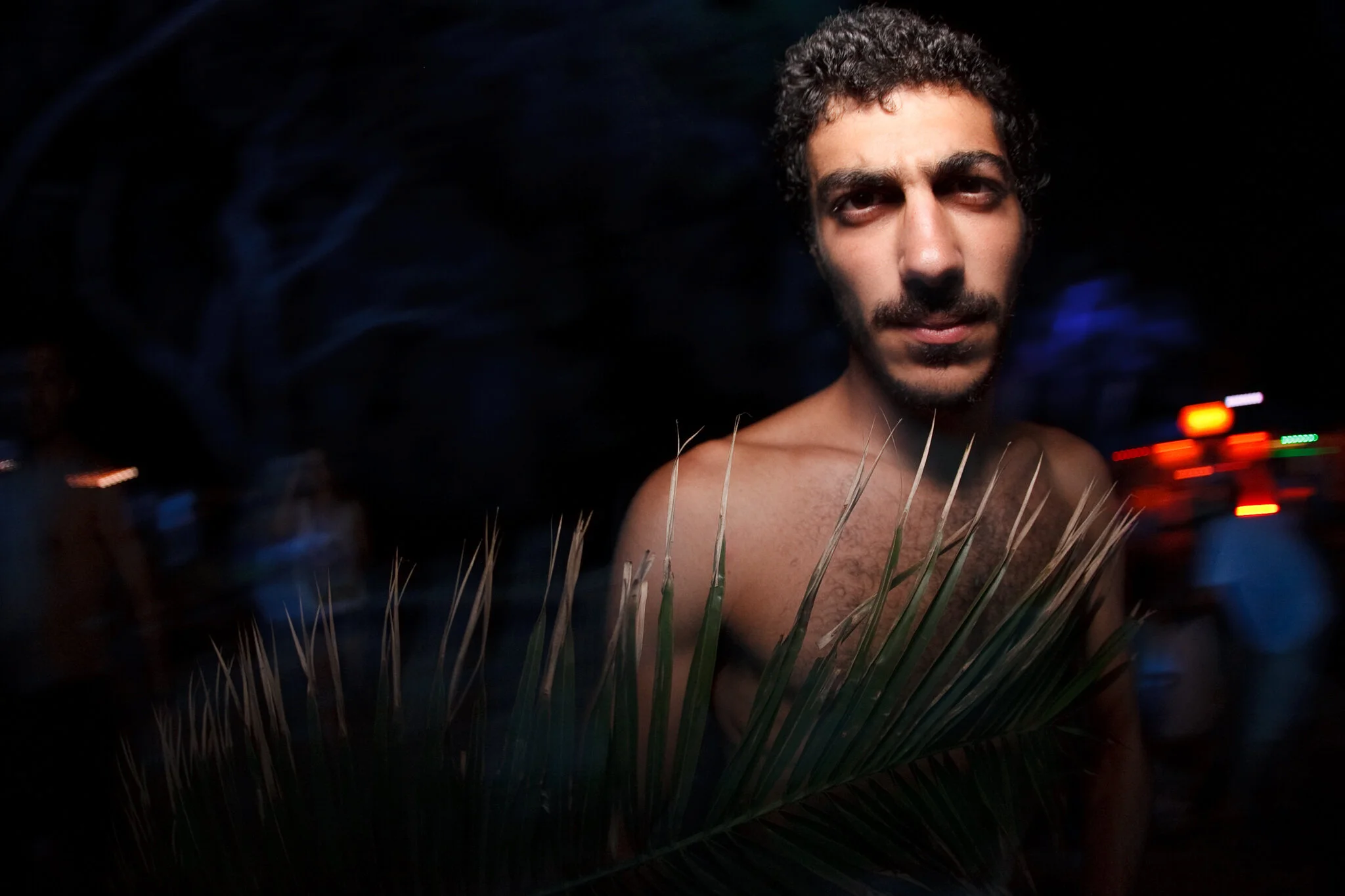 Junger Mann ohne Oberteil, im Dunkeln, blickt in die Kamera, mit Palmblättern im Vordergrund.