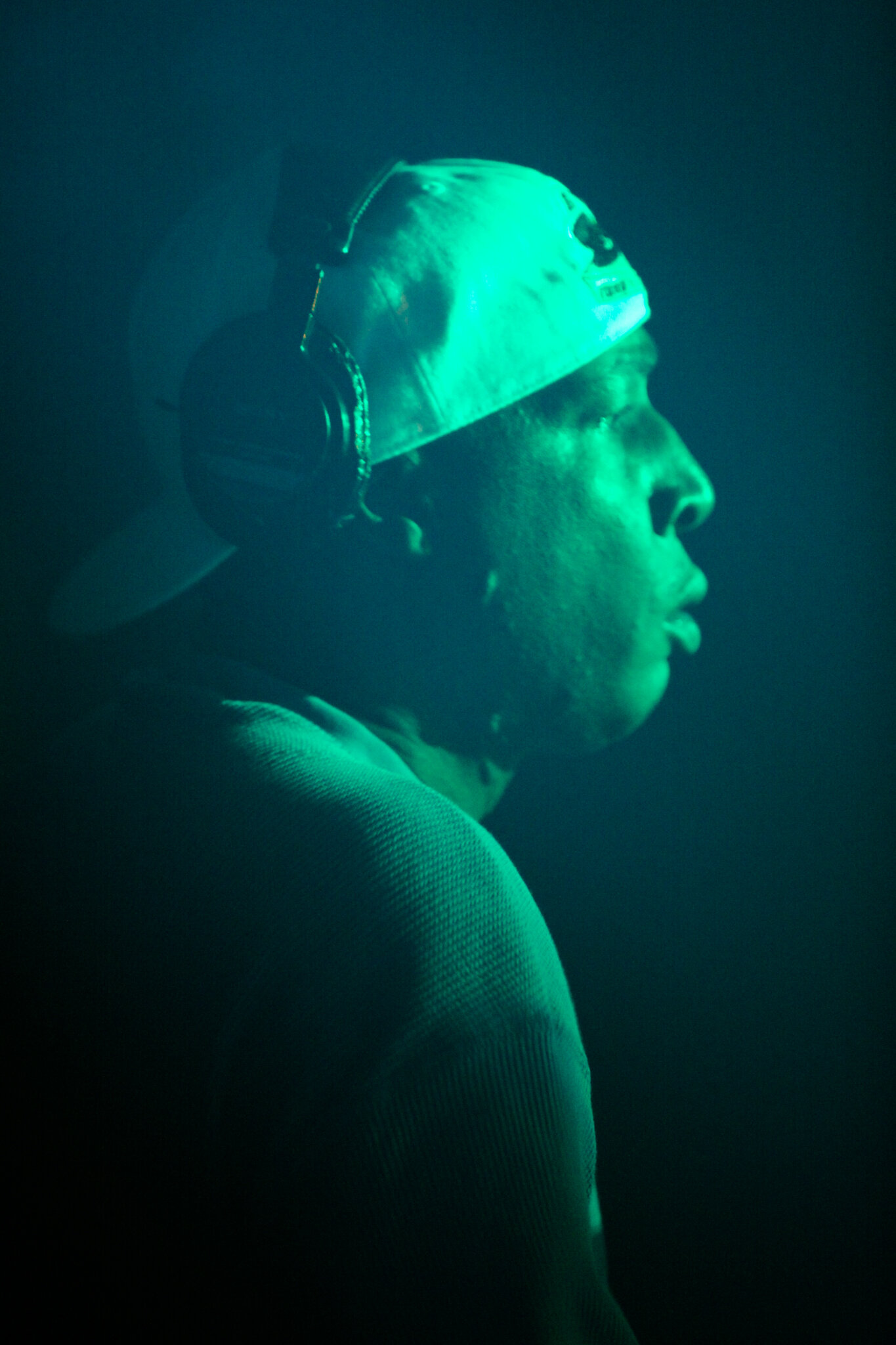 Ein Mann trägt Kopfhörer und einen Schutzhelm, blickt nach rechts in dunklem Licht.