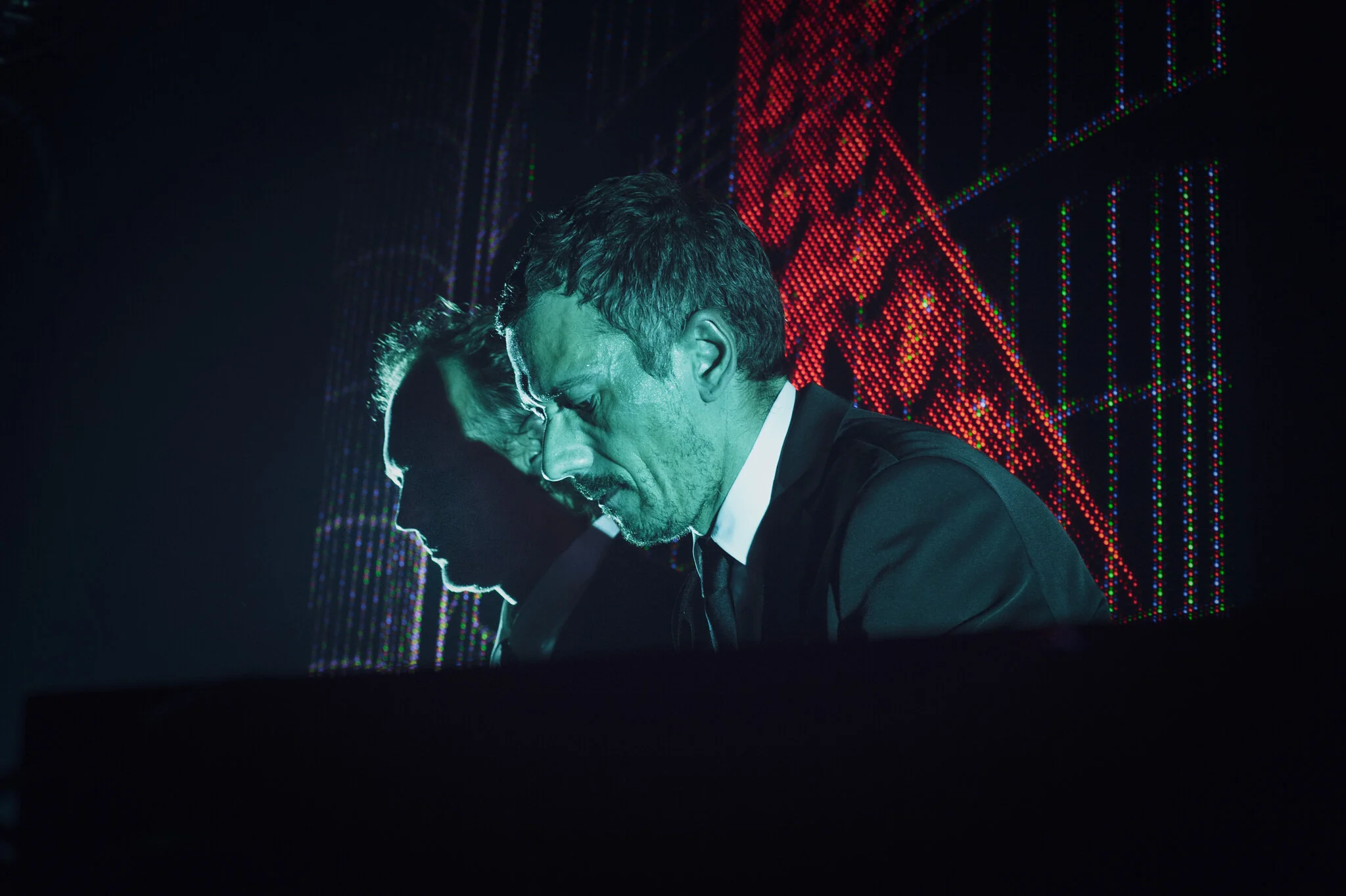 Kruder & Dorfmeister in dunkler Kleidung und Krawatte, die nach vorne gebeugt mit ernster Miene vor einem LED-Wand mit roten und grünen Lichtern stehen.
