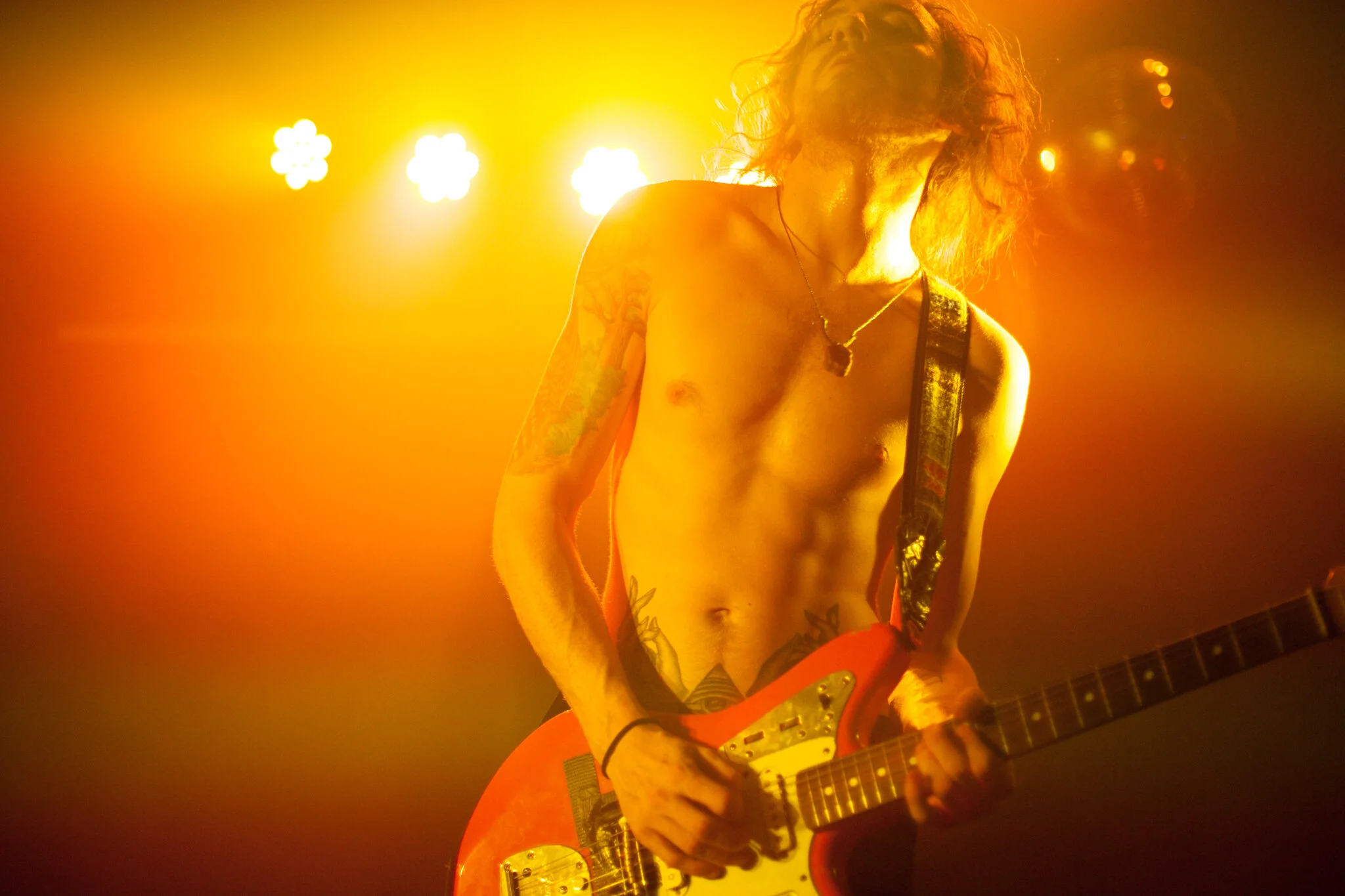 Ein nackter männlicher Rockstar mit Tattoos spielt elektrische Gitarre auf der Bühne, umgeben von gelbem und orangem Bühnenbeleuchtung.