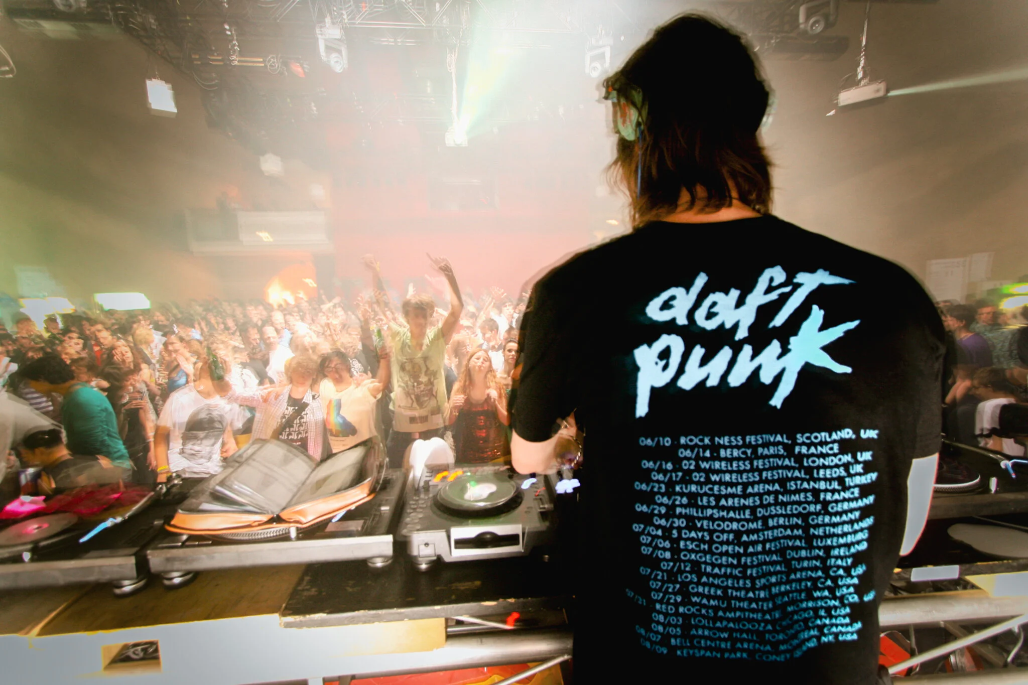 Piedro Winter alias Busy P mit einem ’Daft Punk’ T-Shirt spielt auf einer Party vor einer großen, tanzenden Menschenmenge in einem Club. Die Atmosphäre ist energiegeladen mit bunten Lichtern und Rauch.