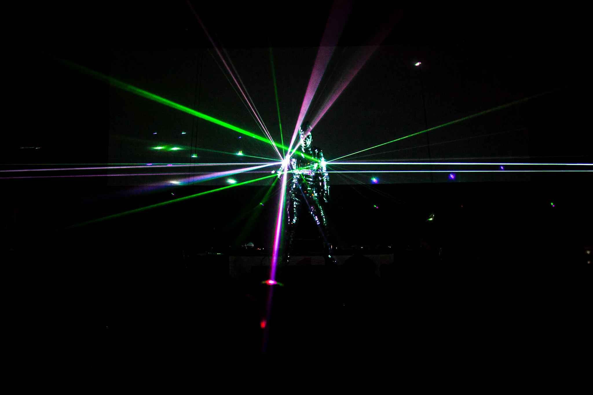 Künstler auf einer Bühne bei einer Lasershow, beleuchtet von grünen, rosa und blauen Laserstrahlen in einem dunklen Raum.