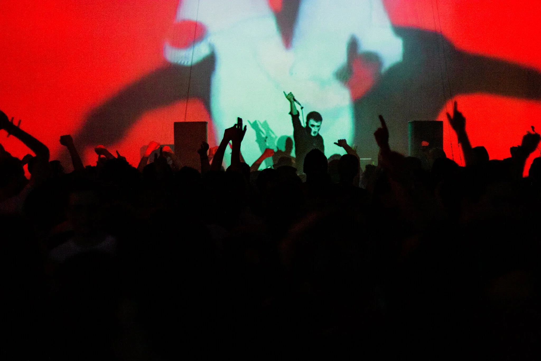 Eine Person auf einer Bühne bei einer Veranstaltung, während im Hintergrund eine große Leinwand mit einem projizierten Bild zu sehen ist, und das Publikum im Vordergrund mit erhobenen Händen.