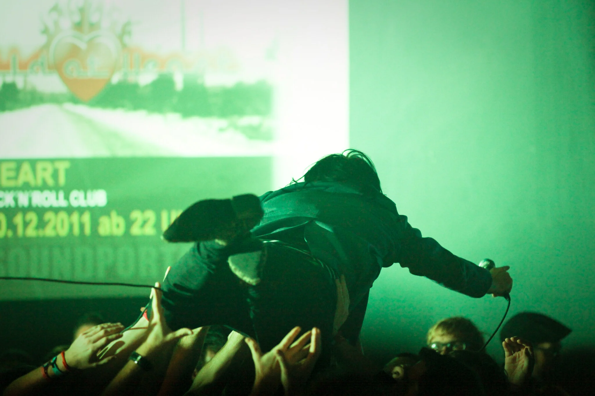 Ein Musiker bei einem Live-Konzert, der auf die Menschen masht, die ihn an der Bühne erreichen. Im Hintergrund eine grüne Lichtprojektion.