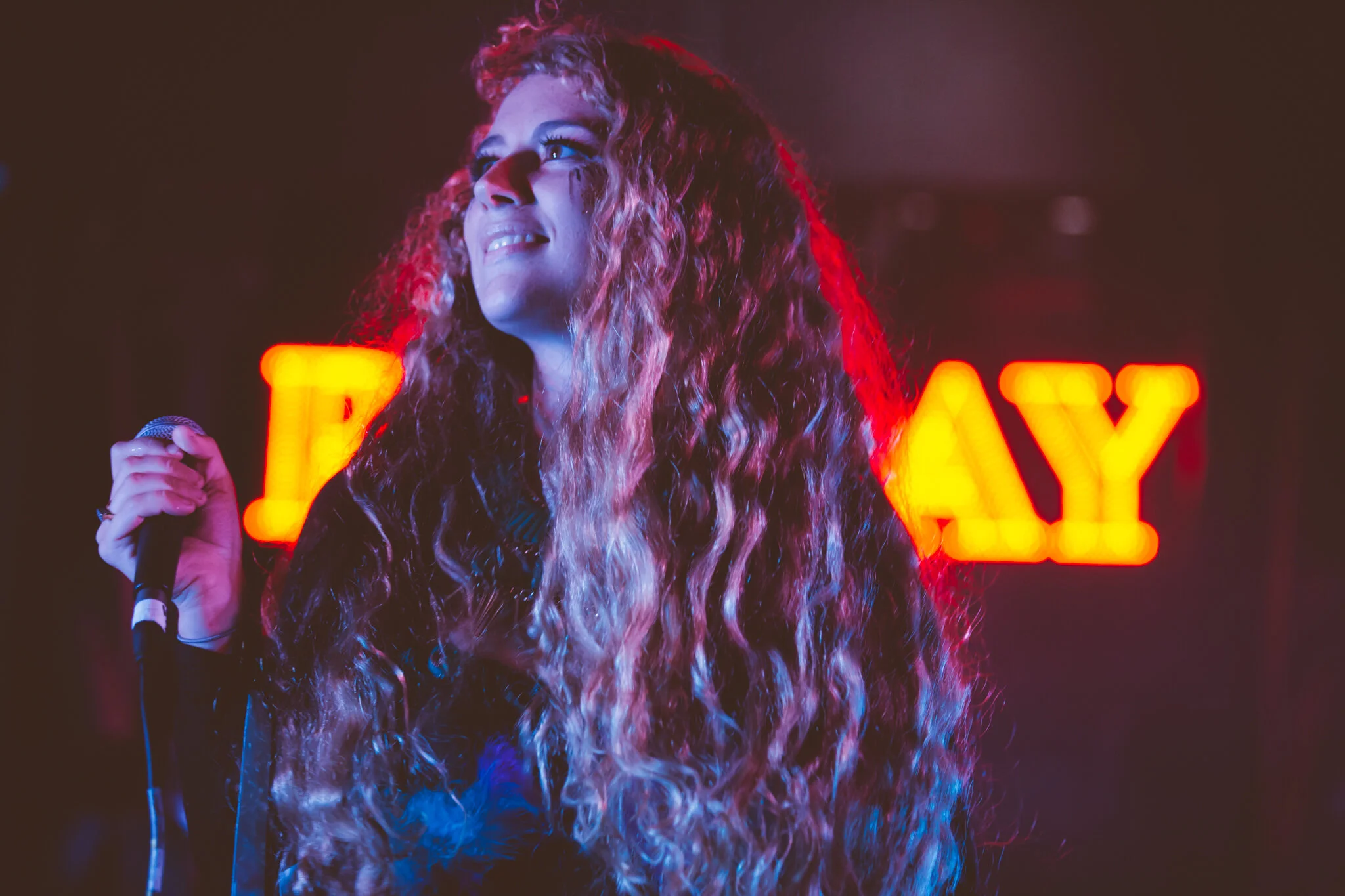 Eine Frau mit langen, lockigen Haaren steht auf einer Bühne, hält ein Mikrofon in der Hand und schaut nach oben. Im Hintergrund leuchtet eine rote Neonreklame, die das Wort 'PLAY' zeigt.