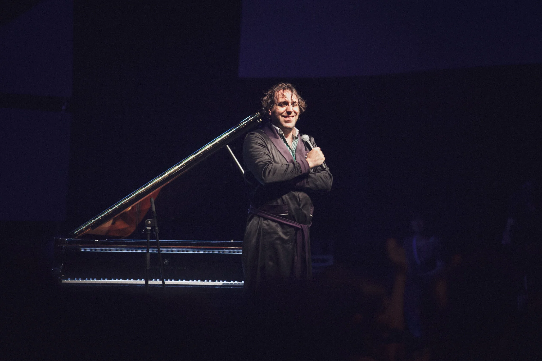 Chilly Gonzales steht auf einer Bühne neben einem Klavier und hält ein Mikrofon, während er spricht oder performt, im Hintergrund dunkler Raum mit wenigen Zuschauern.