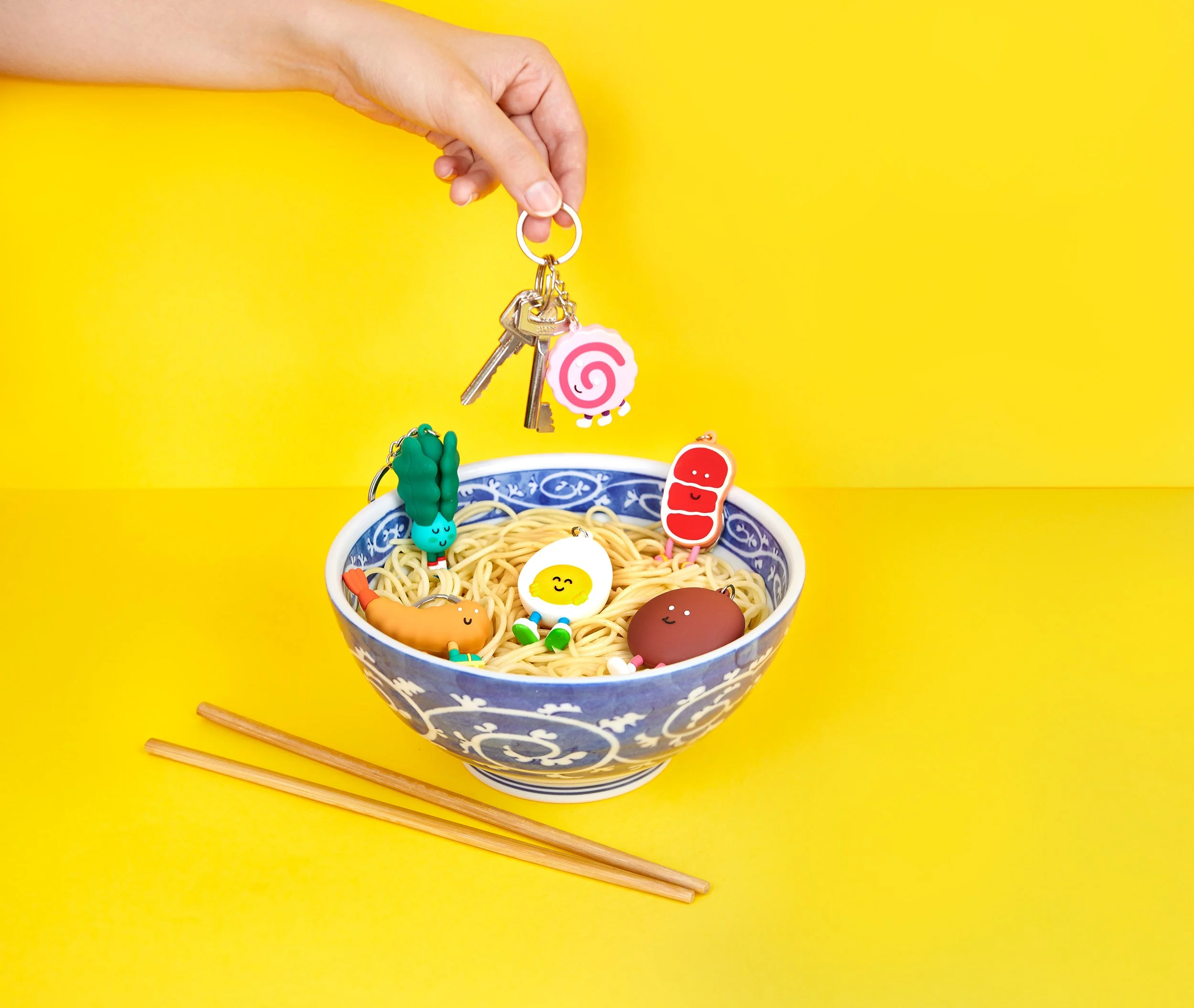 Hand placing kawaii Brosmind ramen ingredient keychains into a noodle bowl on a bright yellow background.