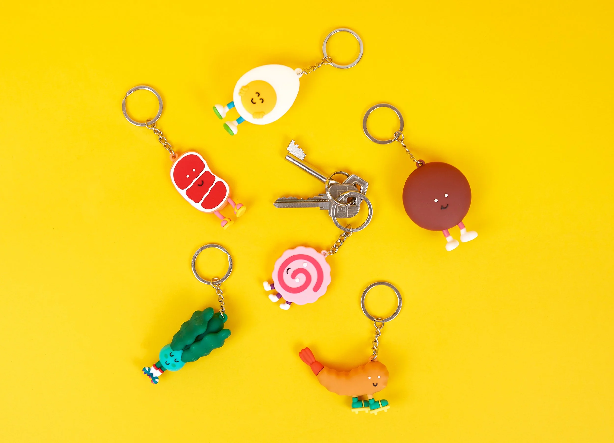 Top-down view of kawaii ramen ingredient keychains attached to metal keys on a yellow background.